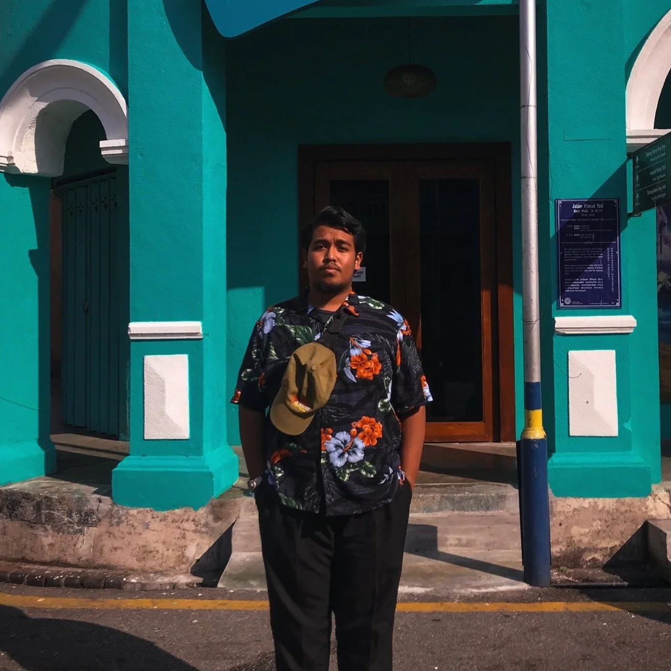 A man standing outside in front of a turquoise building with a wooden door. He is wearing a black Hawaiian shirt with colorful floral patterns, black pants, and has a tan baseball cap hanging from his shirt pocket. The man has short black hair and a beard, and appears to be looking at the camera. The sunlight casts shadows on the scene.