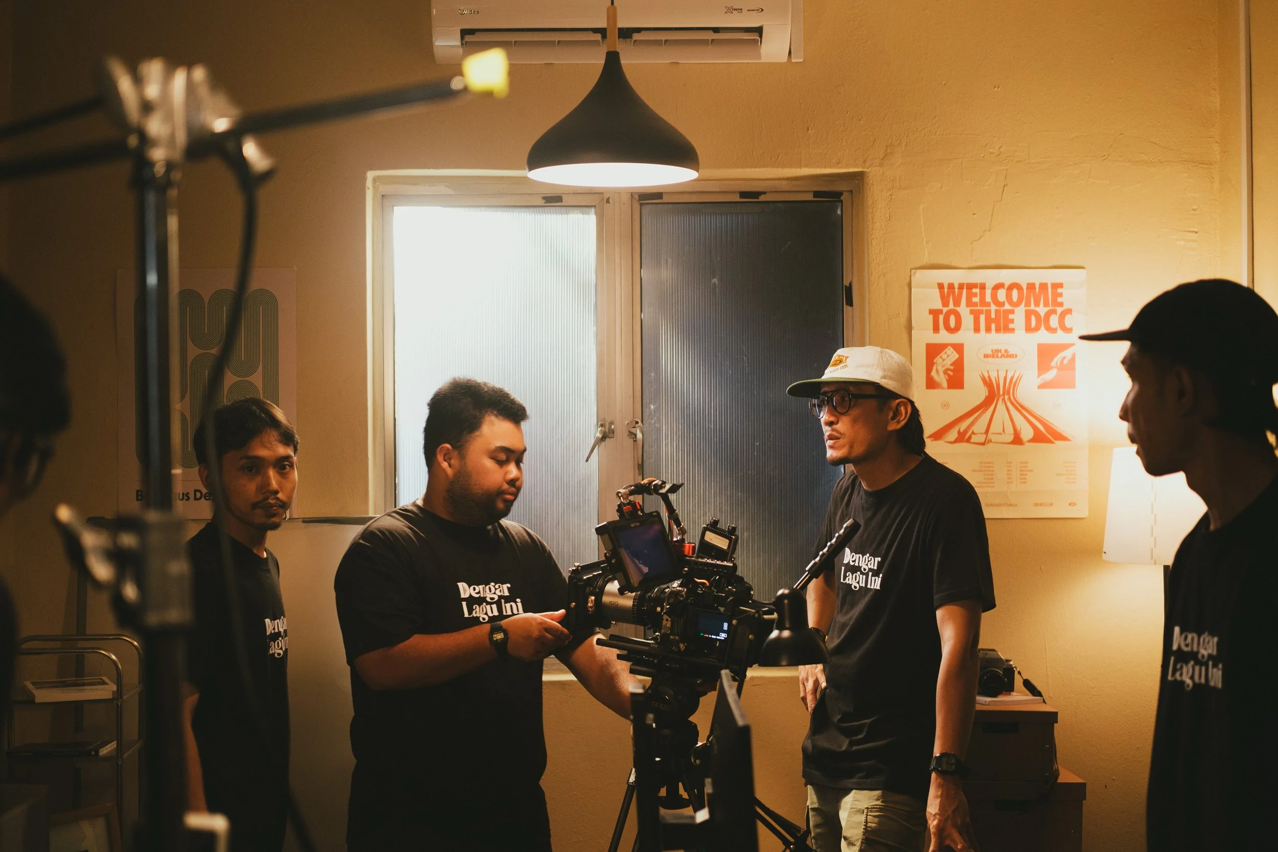A film crew is working in a small room with yellow walls and a window. There are four men, some wearing black shirts with white text. One man is operating a camera, and the others are watching or preparing. A poster on the wall says "Welcome to the D