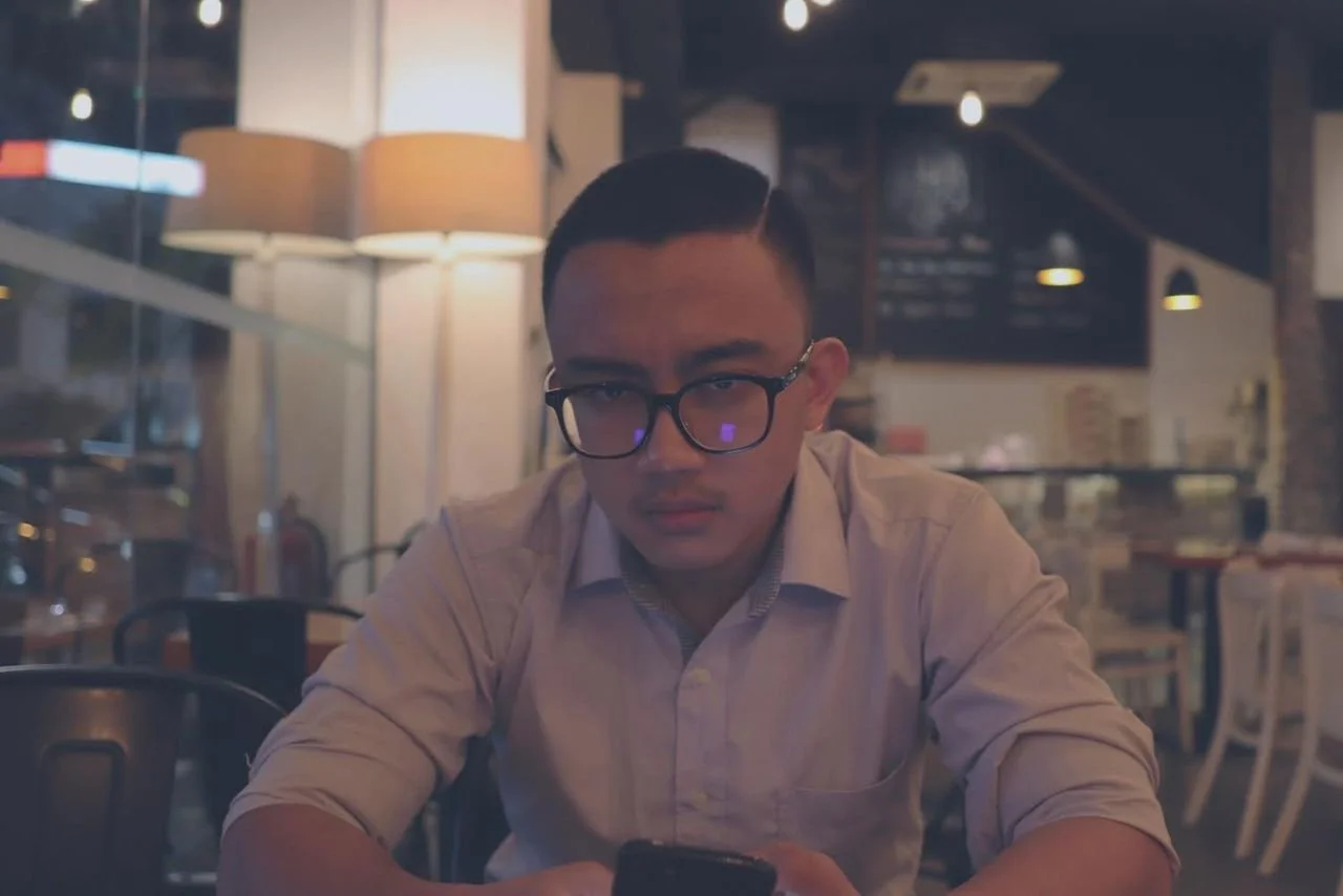 Young man in glasses looking at his phone inside a restaurant or cafe with modern decor and warm lighting.