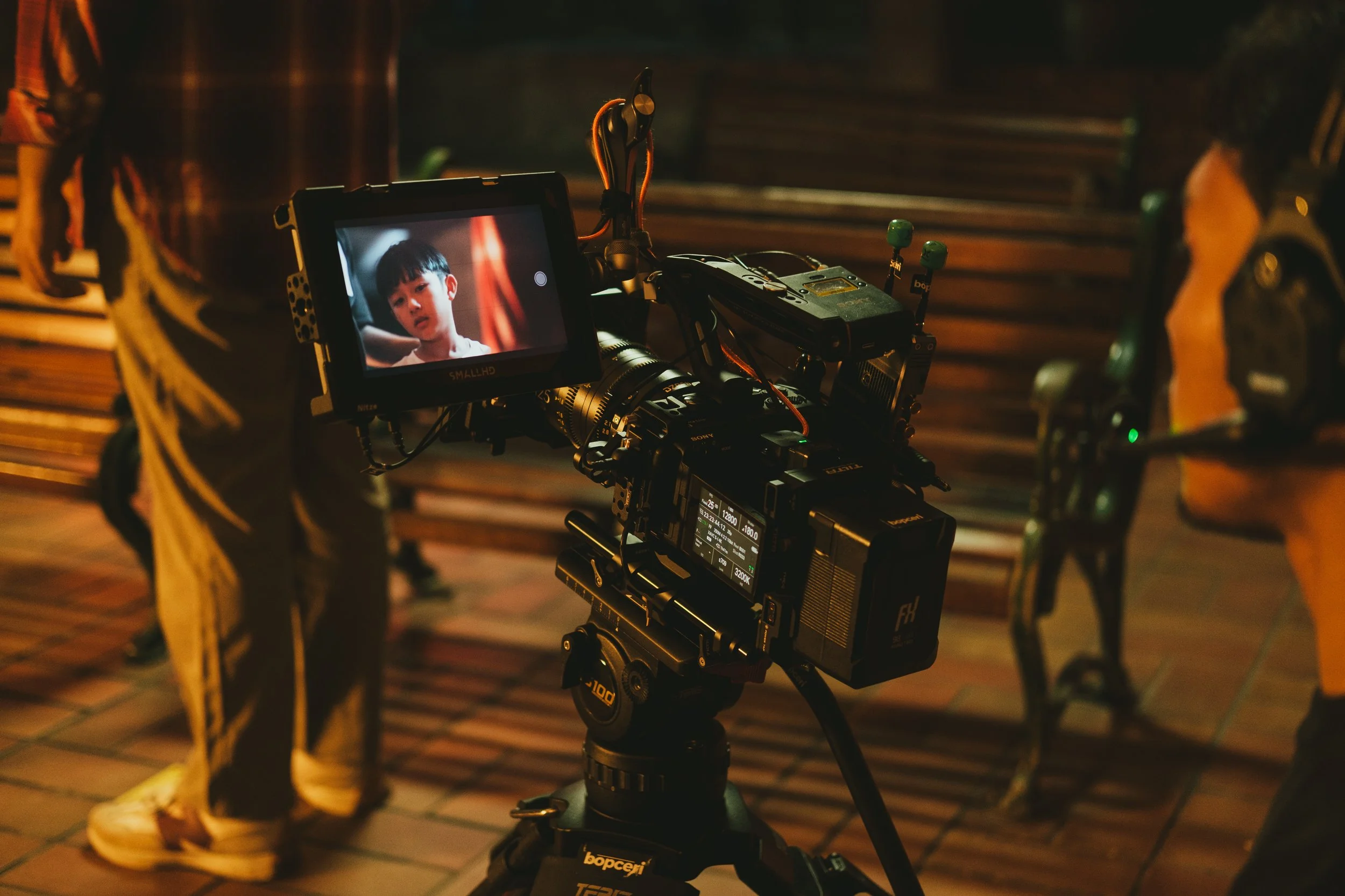 A camera on a tripod filming a young boy with a bowl haircut, who is looking toward the camera, in an indoor setting with wooden benches in the background.