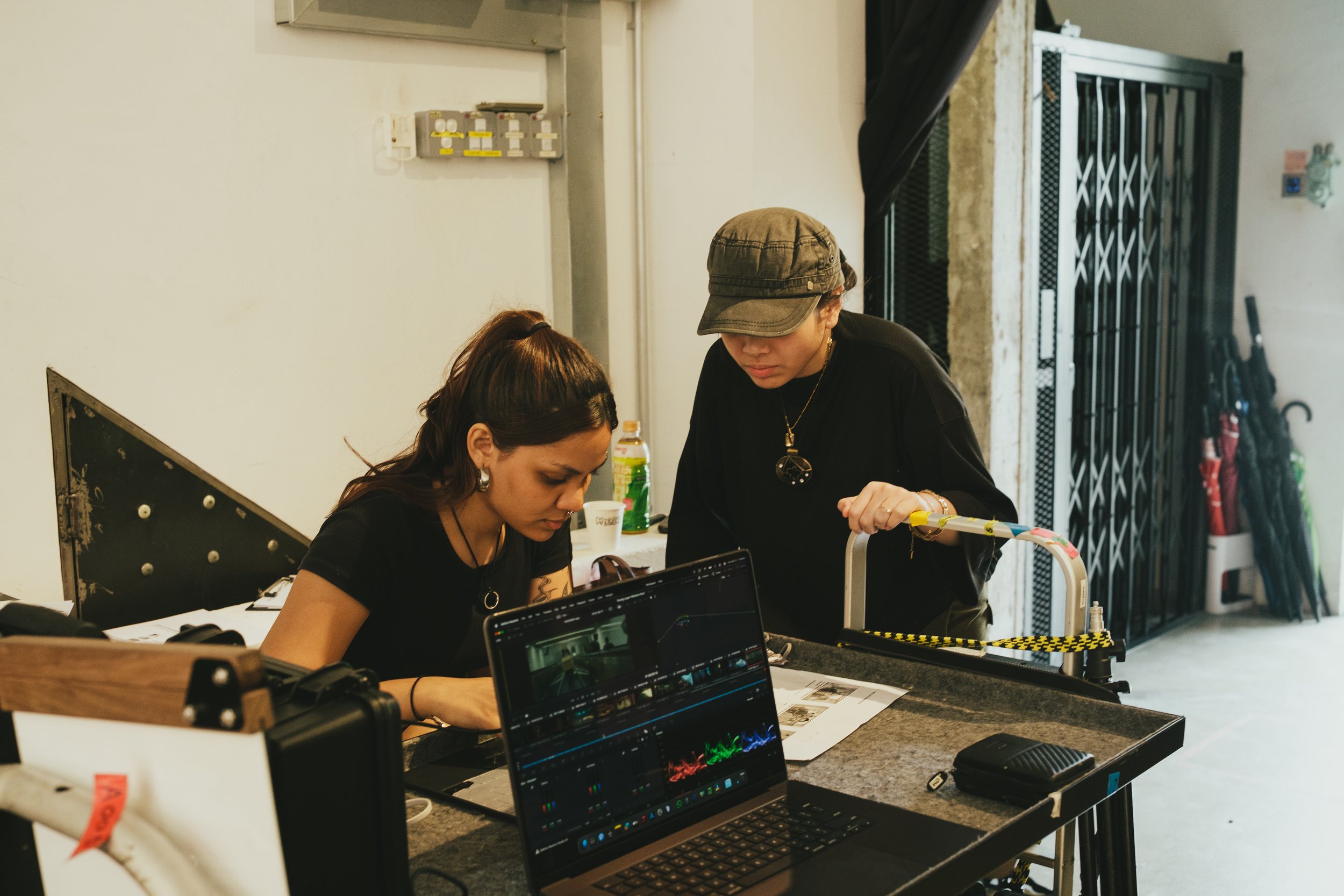 Two women working together at a table with a laptop, a printer, and various papers, in a room with a black gate and umbrellas in the background.
