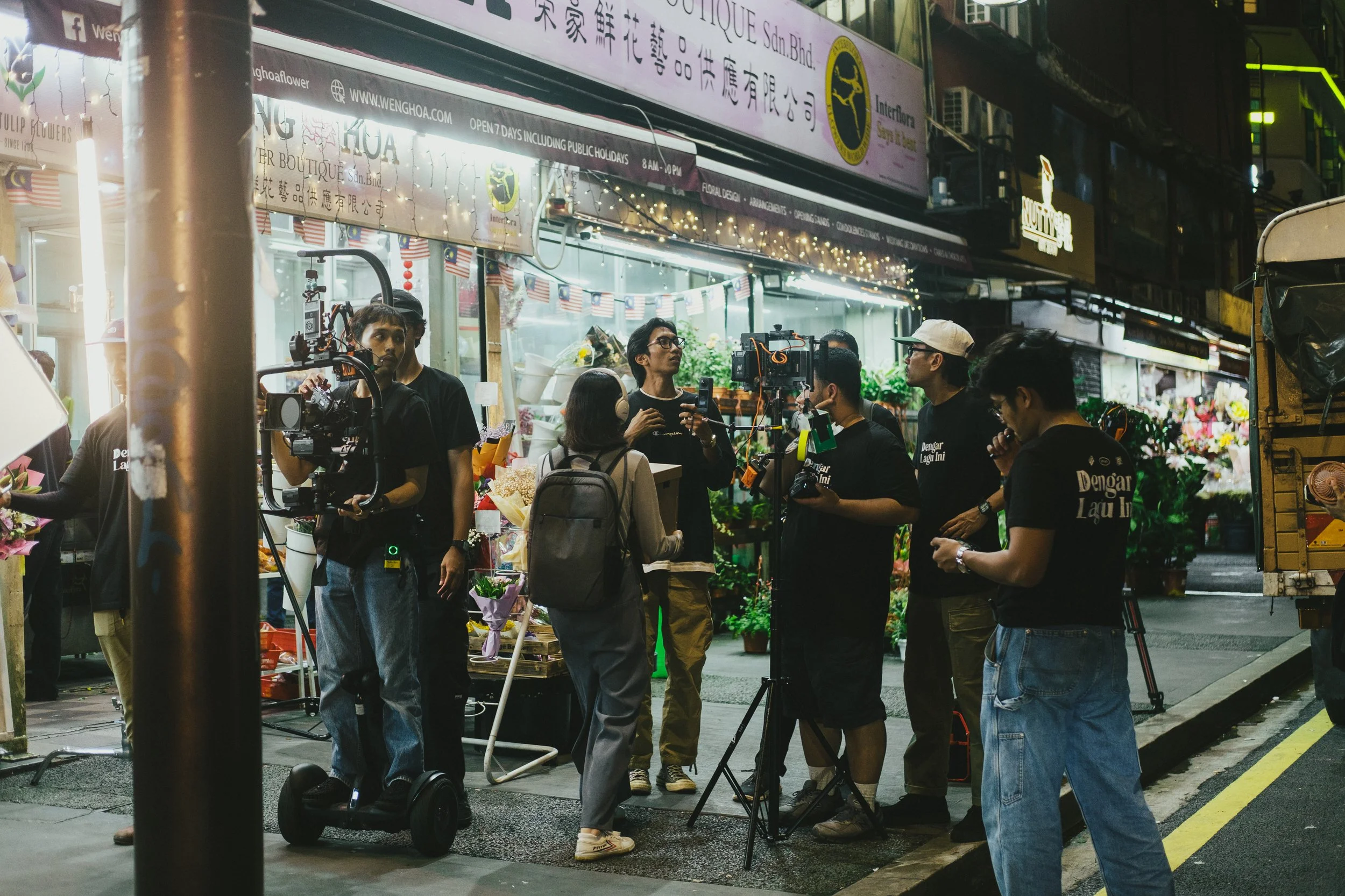A group of people, including a woman with a backpack, are filming or broadcasting live outside a flower shop at night. They are surrounded by camera equipment, and the shop is decorated with string lights and flowers.