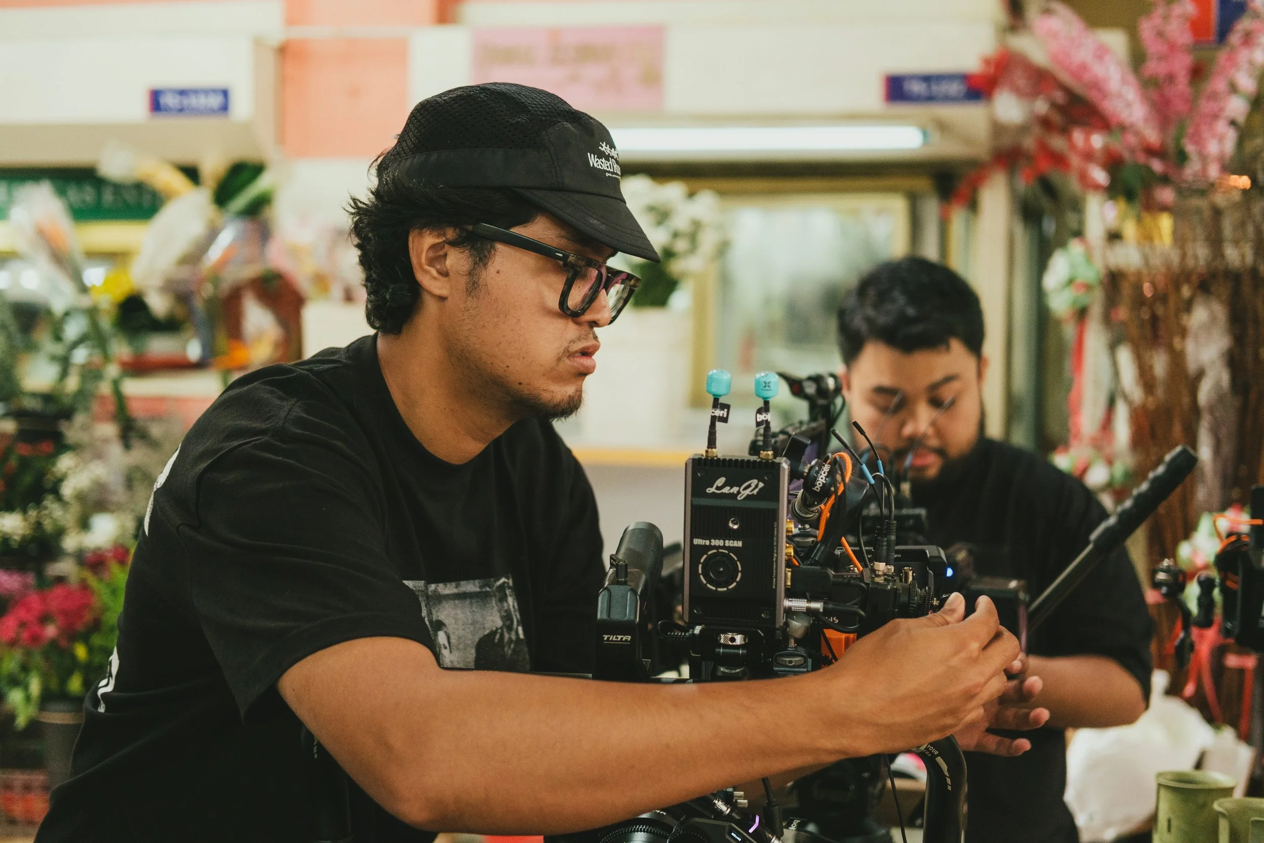 Two men working on a film or video camera in a flower shop or florist's shop.