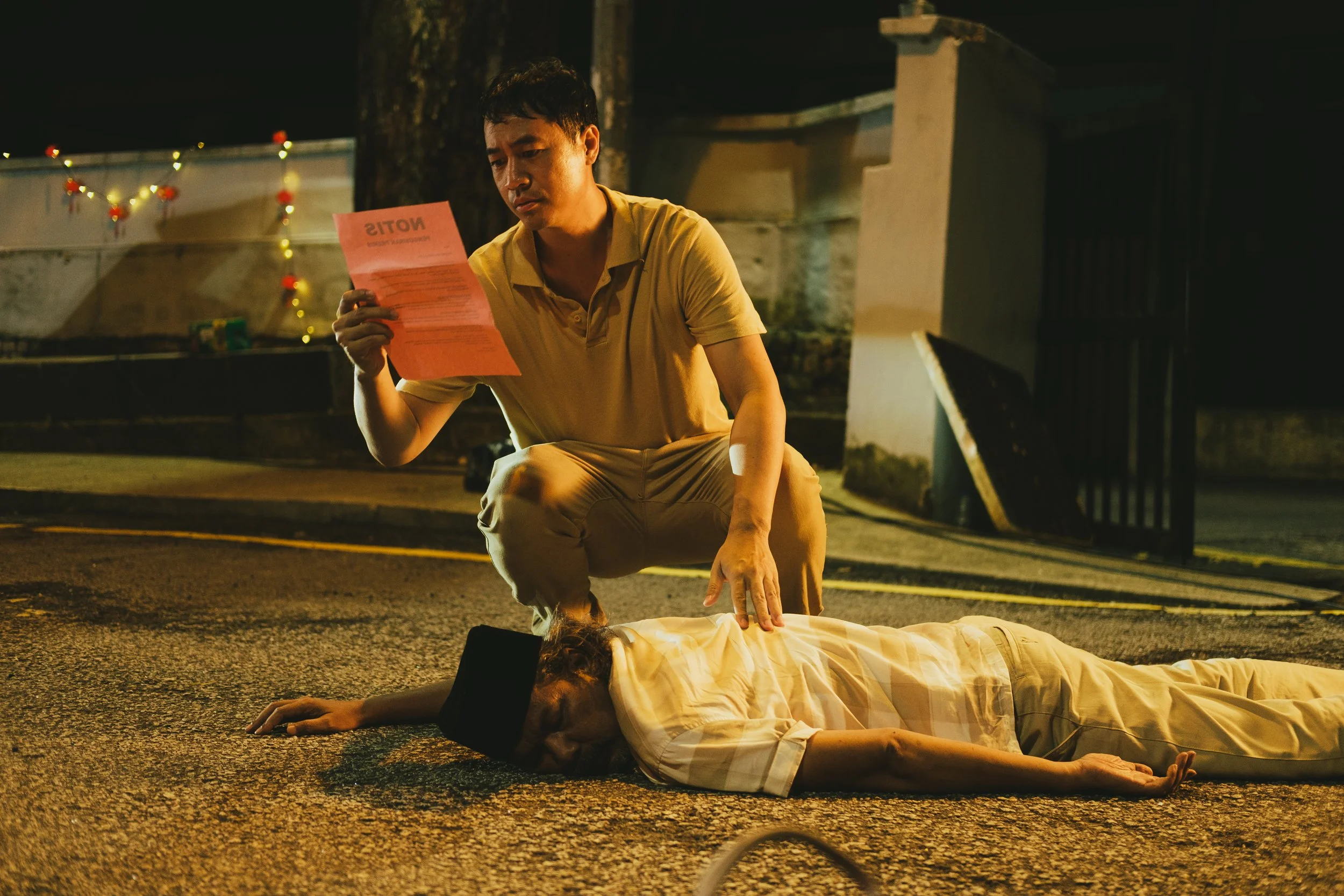 A young man kneels beside a woman lying unconscious on the street at night. The man is holding a pink sheet of paper and appears to be distressed, while the woman is on her side with a black cloth over her face. There are Christmas lights and decorat