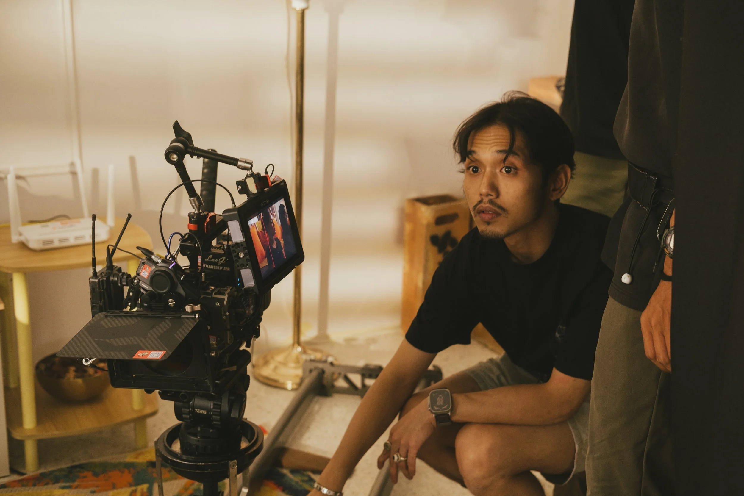 A man crouches near a camera on a tripod, looking at the camera's monitor. Another person stands nearby, partially visible. The setting appears to be indoors with a lamp and some furniture in the background.