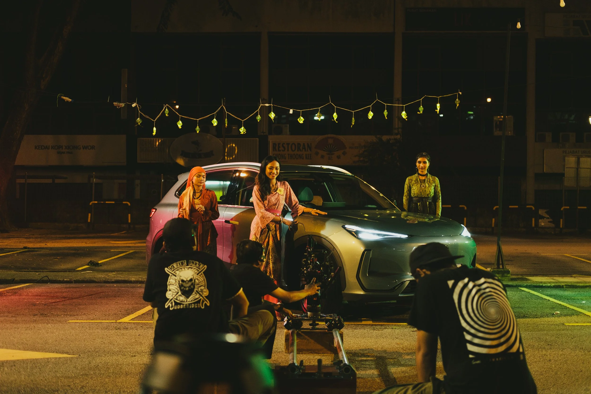 A nighttime photoshoot featuring three women in traditional attire standing next to a modern silver car. The scene is outdoors in a parking lot with filming equipment and crew members present.
