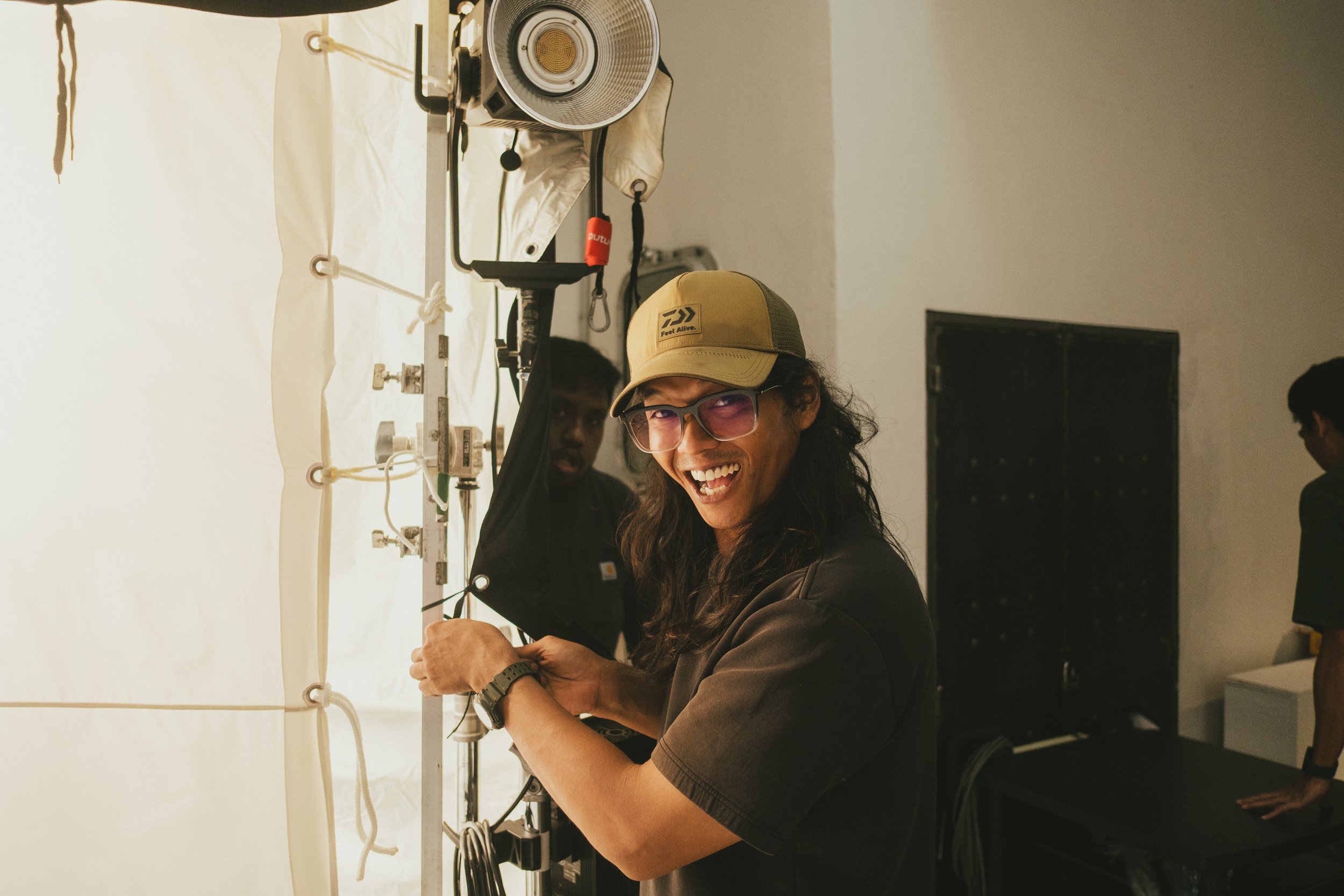 Person smiling with long hair and sunglasses, wearing a beige cap, standing next to photography equipment and a white backdrop, in a studio setting.