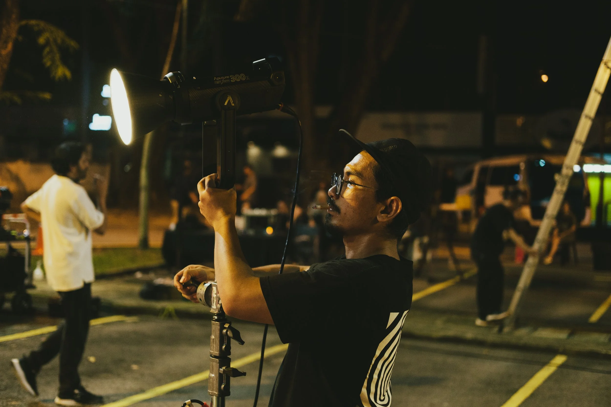 A man operating a large flashlight on a stand at night in a parking lot during a film or photo shoot, with other people and equipment in the background.