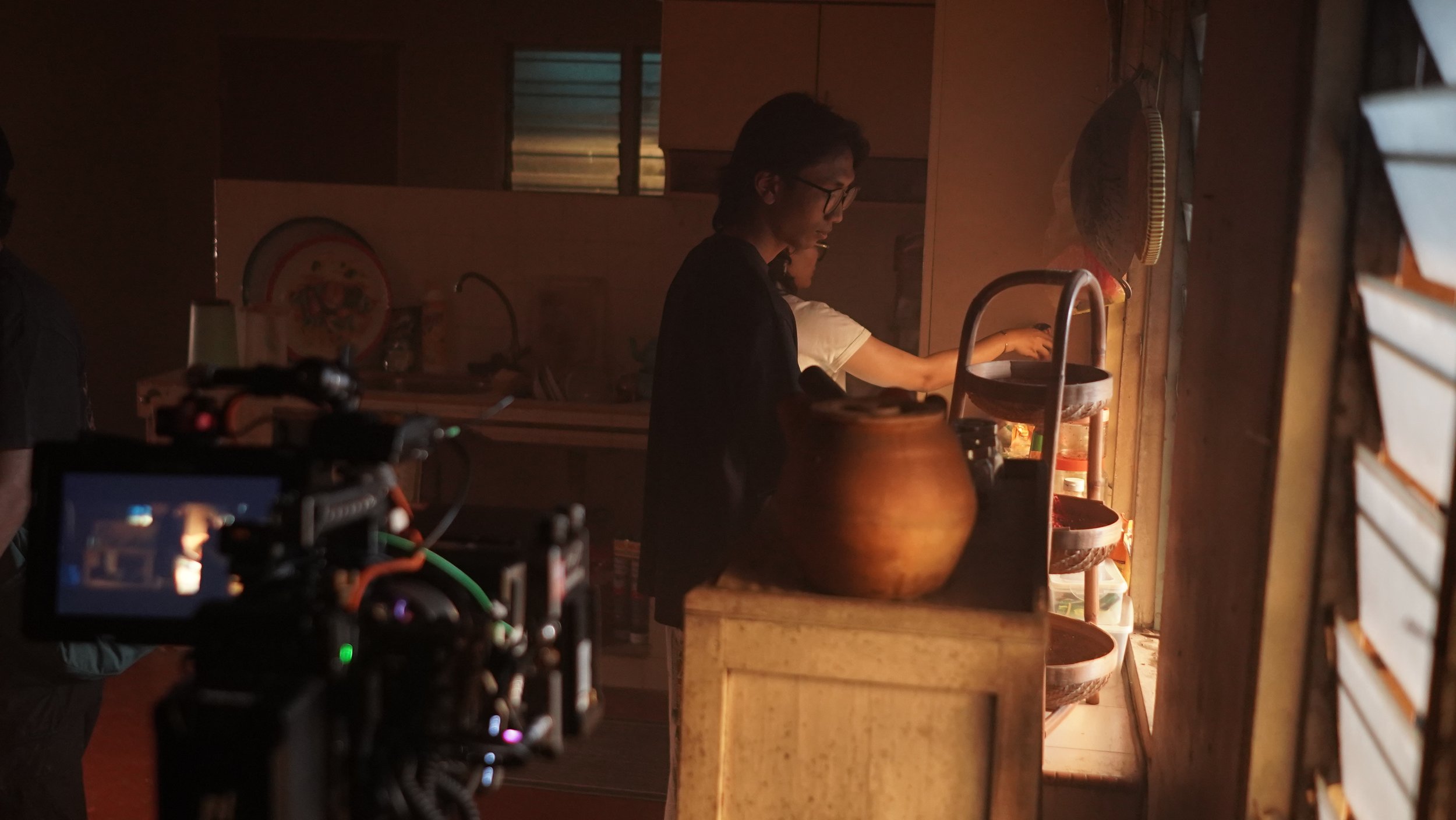 Person standing in a dimly lit kitchen, preparing food or drink, with a camera recording from the foreground.