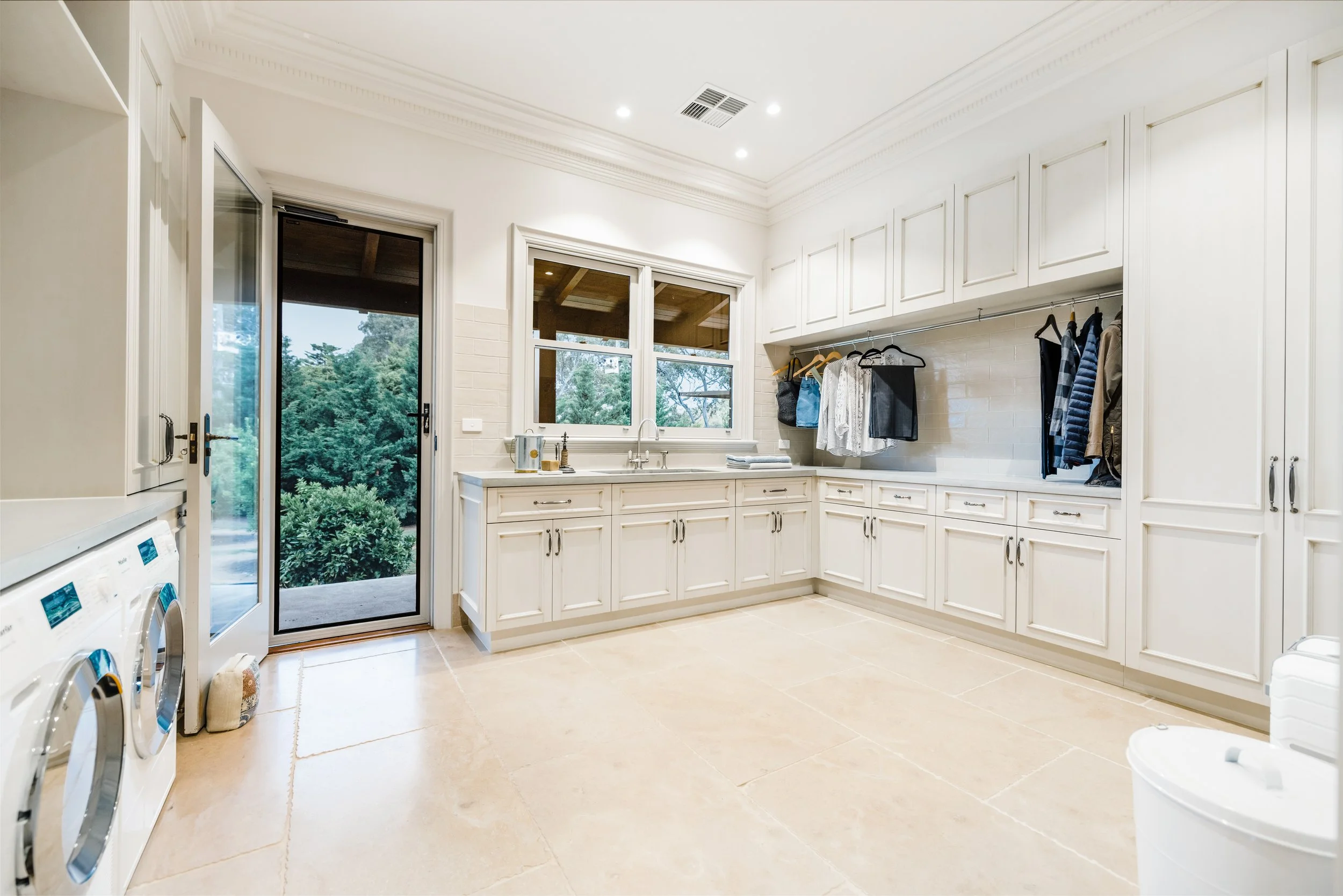 Laundry, cream coloured traditional style cabinetry, hanging rod for clothes, limestone floors