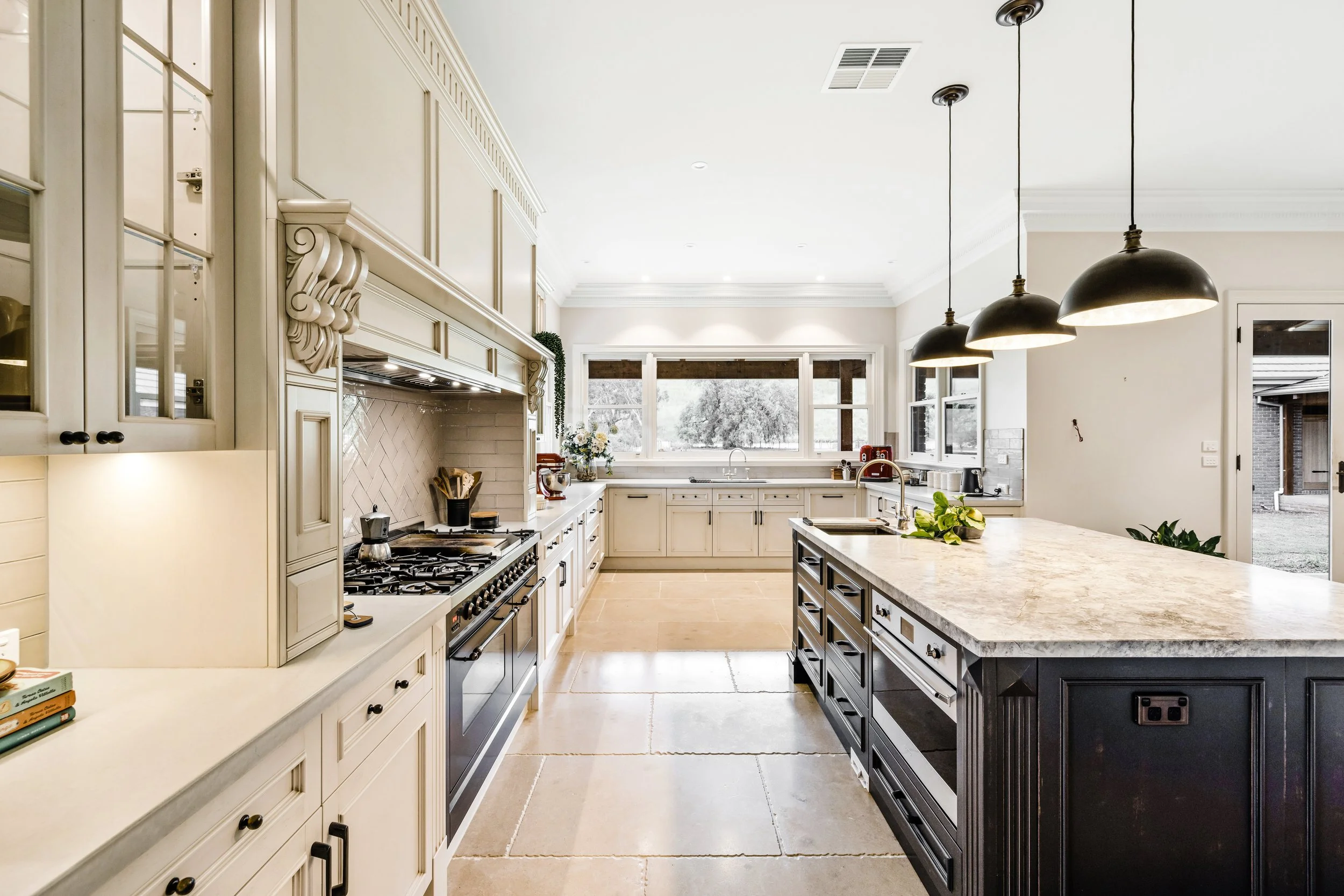 French Country Kitchen, cream coloured cabinetry with black island cabinetry, Dolomite stone island benchtop, surrounding concrete benchtops, chevron tiled splashback, Ilve black 1200mm freestanding cooker, 3 black and gold pendant lights hanging ove