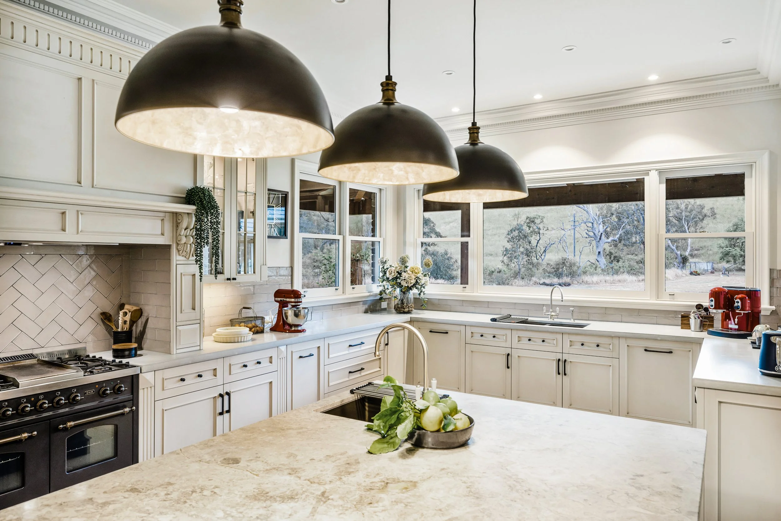 French Country Kitchen, Dolomite stone island benchtop, surrounding concrete benchtops, chevron tiled splashback, Ilve black 1200mm freestanding cooker, 3 black and gold pendant light fixtures hanging over island bench.