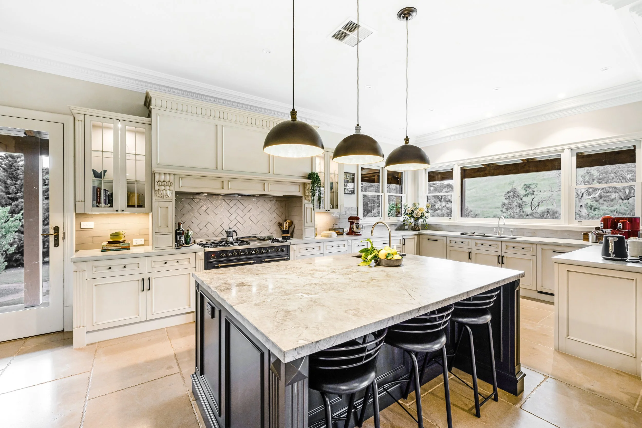 French Country Kitchen, Dolomite stone island benchtop, surrounding concrete benchtops, chevron tiled splashback, Ilve black 1200mm freestanding cooker, 3 black and gold pendant light fixtures hanging over island bench, Limestone Floors