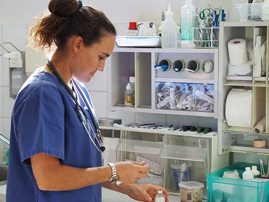 Eine Krankenschwester in blauer Uniform in einem klinischen Raum, die eine Flasche hält, mit medizinischem Zubehör und Medikamenten im Hintergrund.