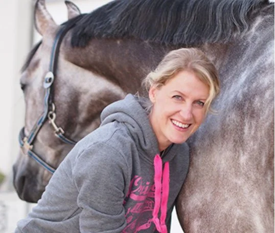 Eine lachende Frau in einem grauen Hoodie steht neben einem Pferd, das einen schwarzen Kopf und graues Fell hat.