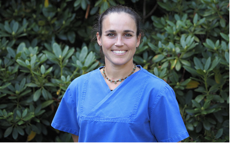 Eine Frau in blauer medizinischer Kleidung steht vor grünen Laubbäumen und lächelt.