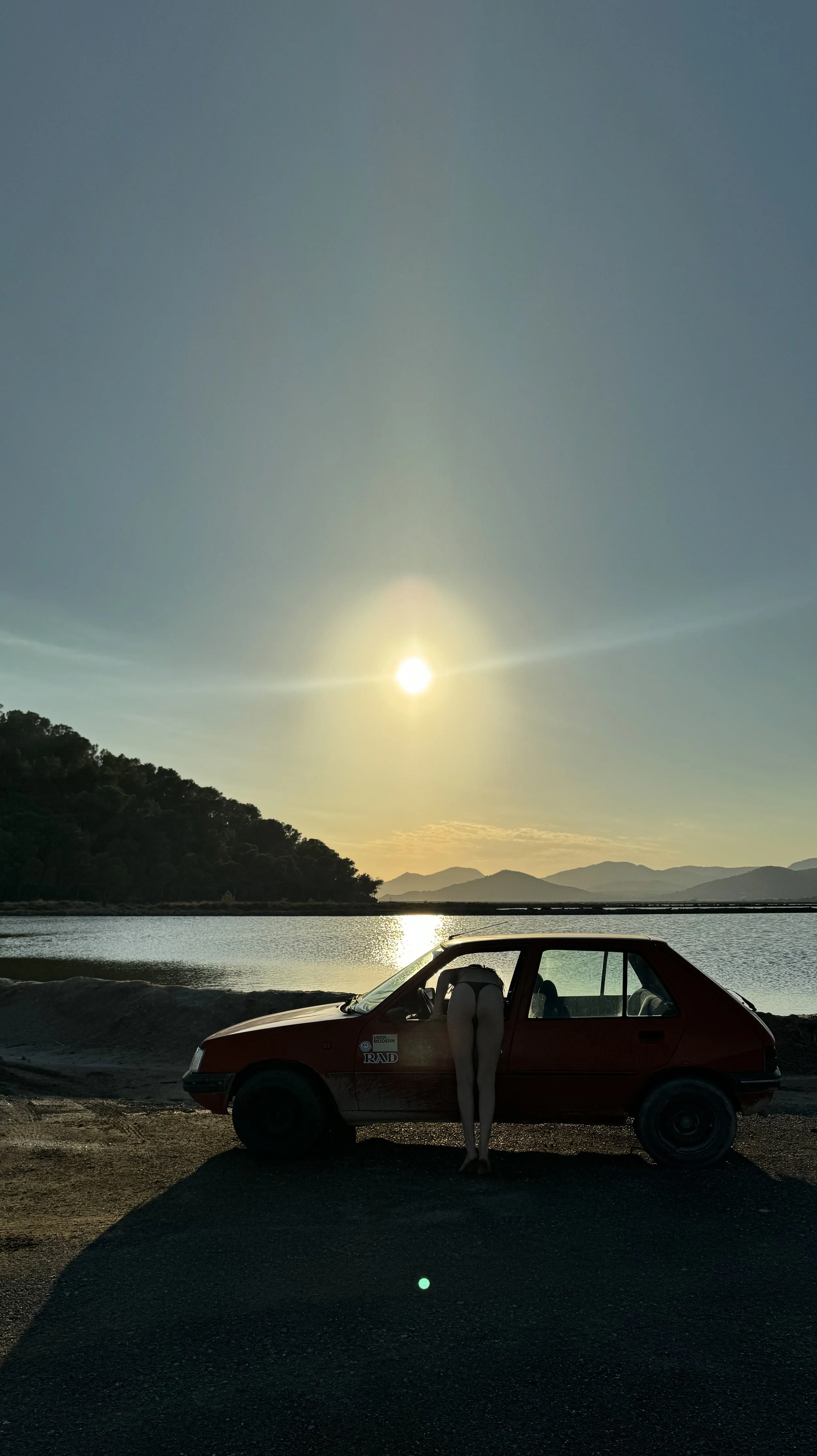 A photo of a landscape during sunset or sunrise with a lake, mountains, and trees in the background. In the foreground, there is a red car with a woman's legs hanging out of the driver side door.