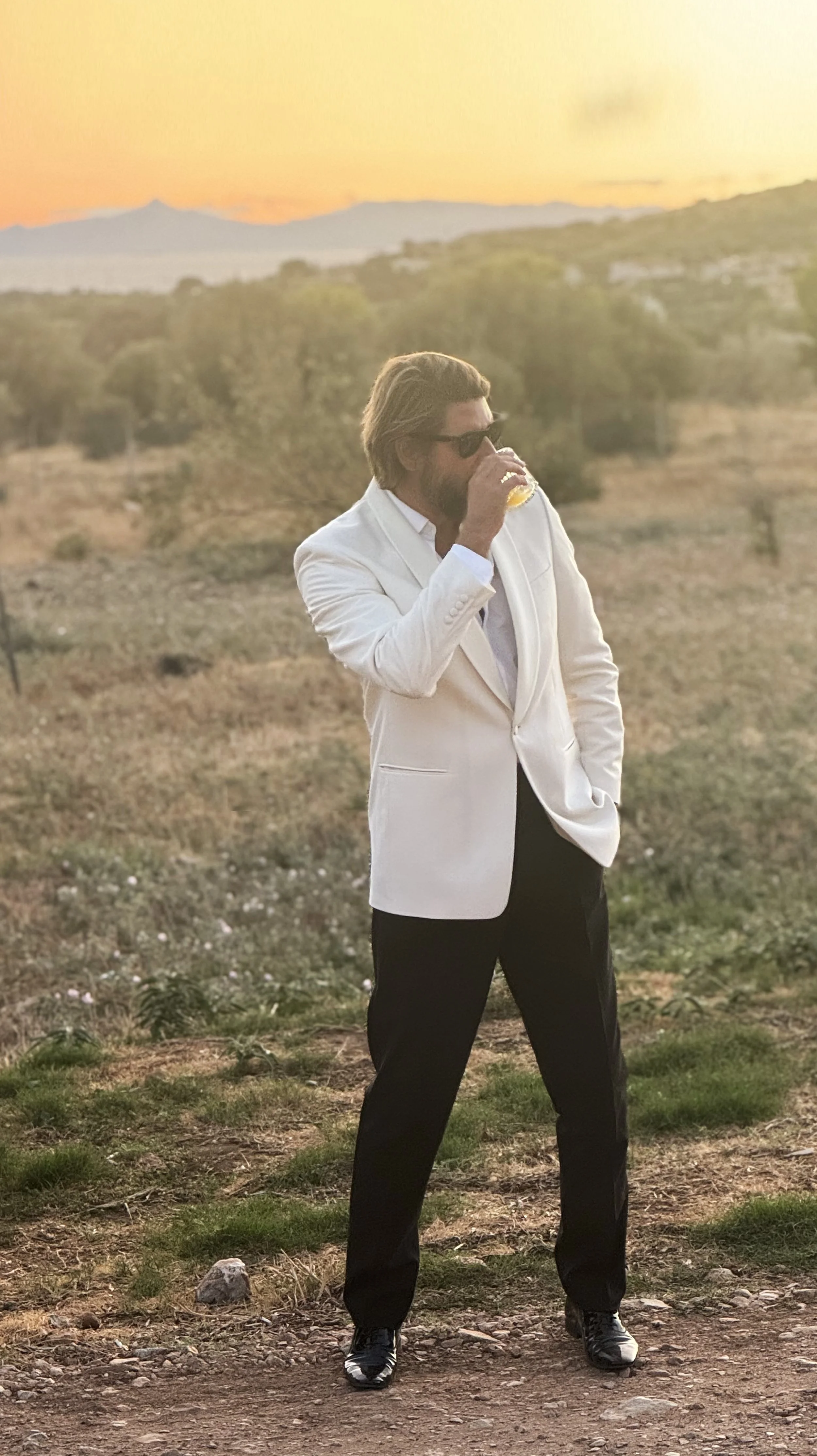 Man with long hair, beard, sunglasses, wearing white blazer and black pants, standing outdoors at sunset, drinking from a glass.
