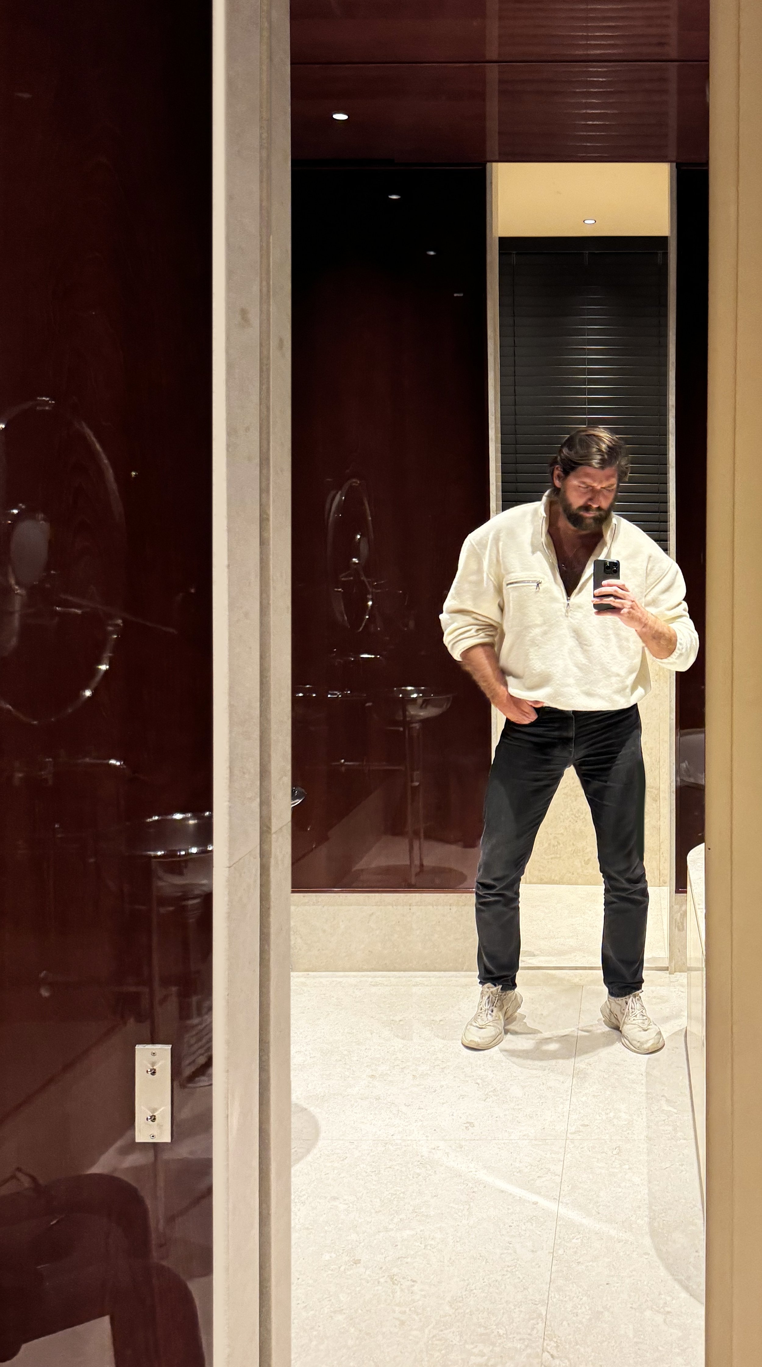 Man taking a mirror selfie in a fitting room, wearing a white fleece jacket, black pants, and white sneakers.