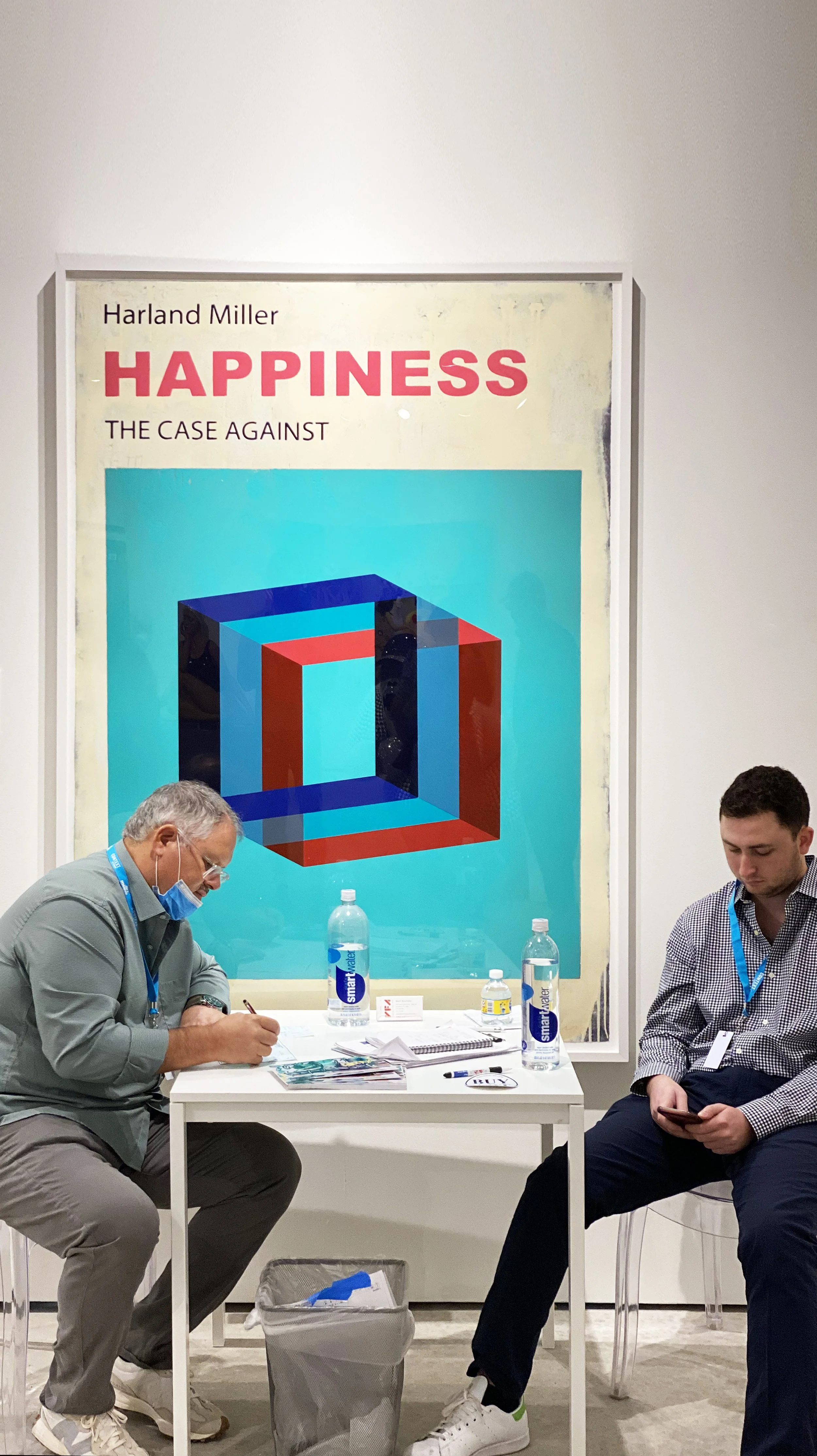 Two men sitting at a small white table with water bottles, papers, and pens, in front of a large poster for a book titled 'Happiness: The Case Against' by Harland Miller, featuring a colorful geometric cube design.