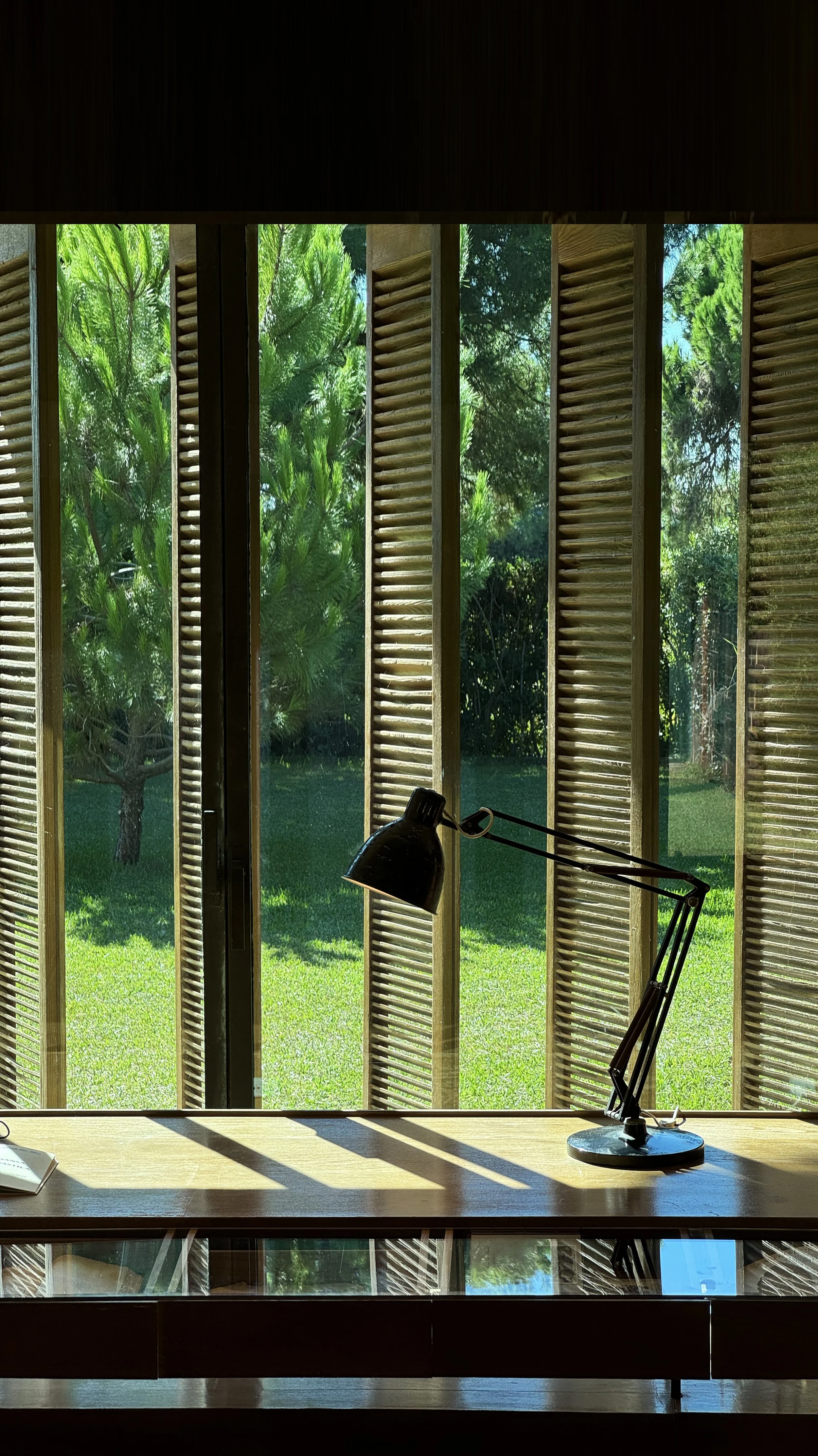 Benjamin Liatoud designs bespoke luxury architecture worldwide.View from inside of a desk with a black adjustable desk lamp, looking out through vertical wooden window shutters to a green outdoor garden with trees and grass.