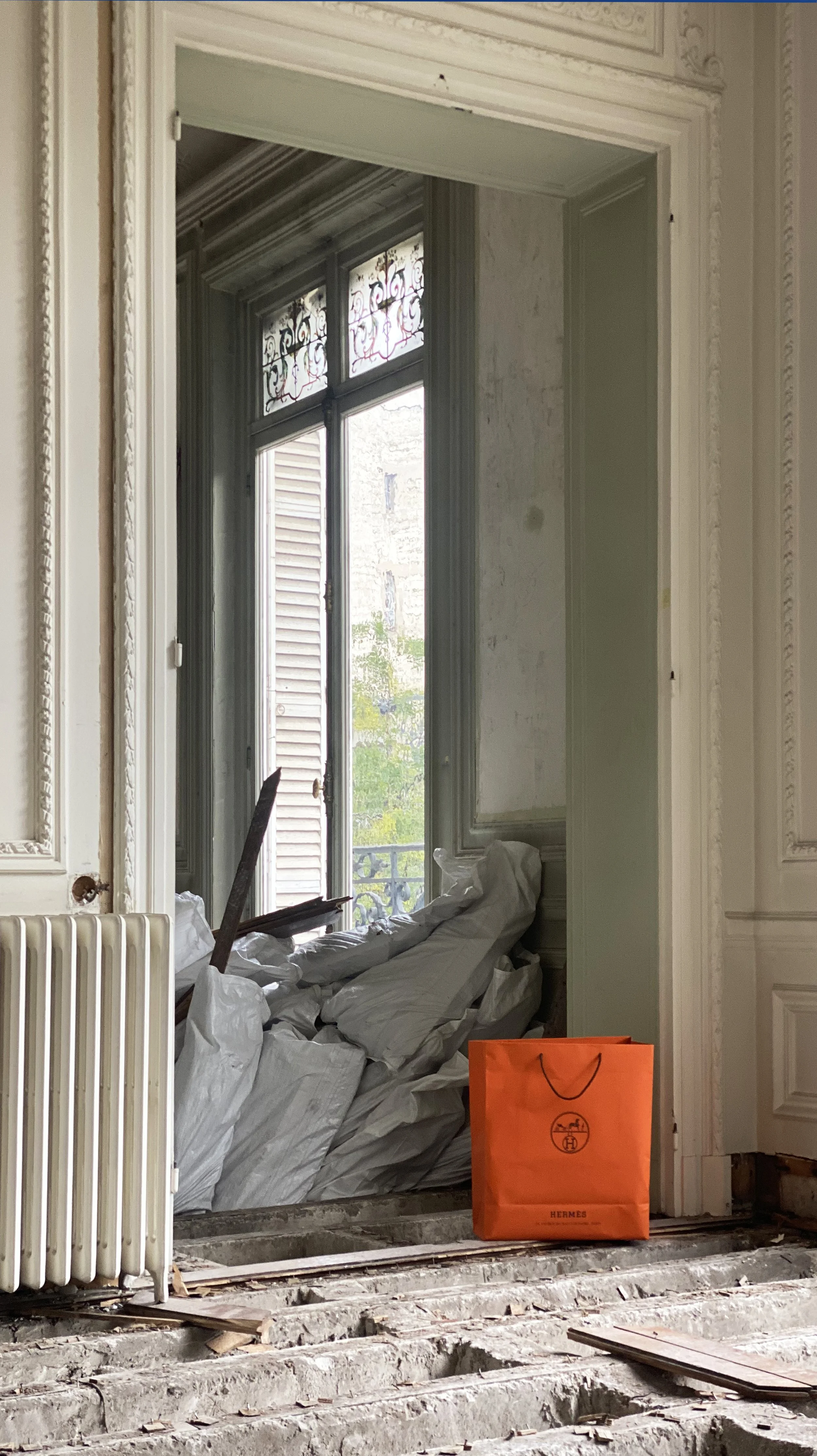 Room undergoing renovation with debris and white bags piled near a large window with stained glass panels, an orange Hermès shopping bag on the floor, and exposed wooden floorboards.