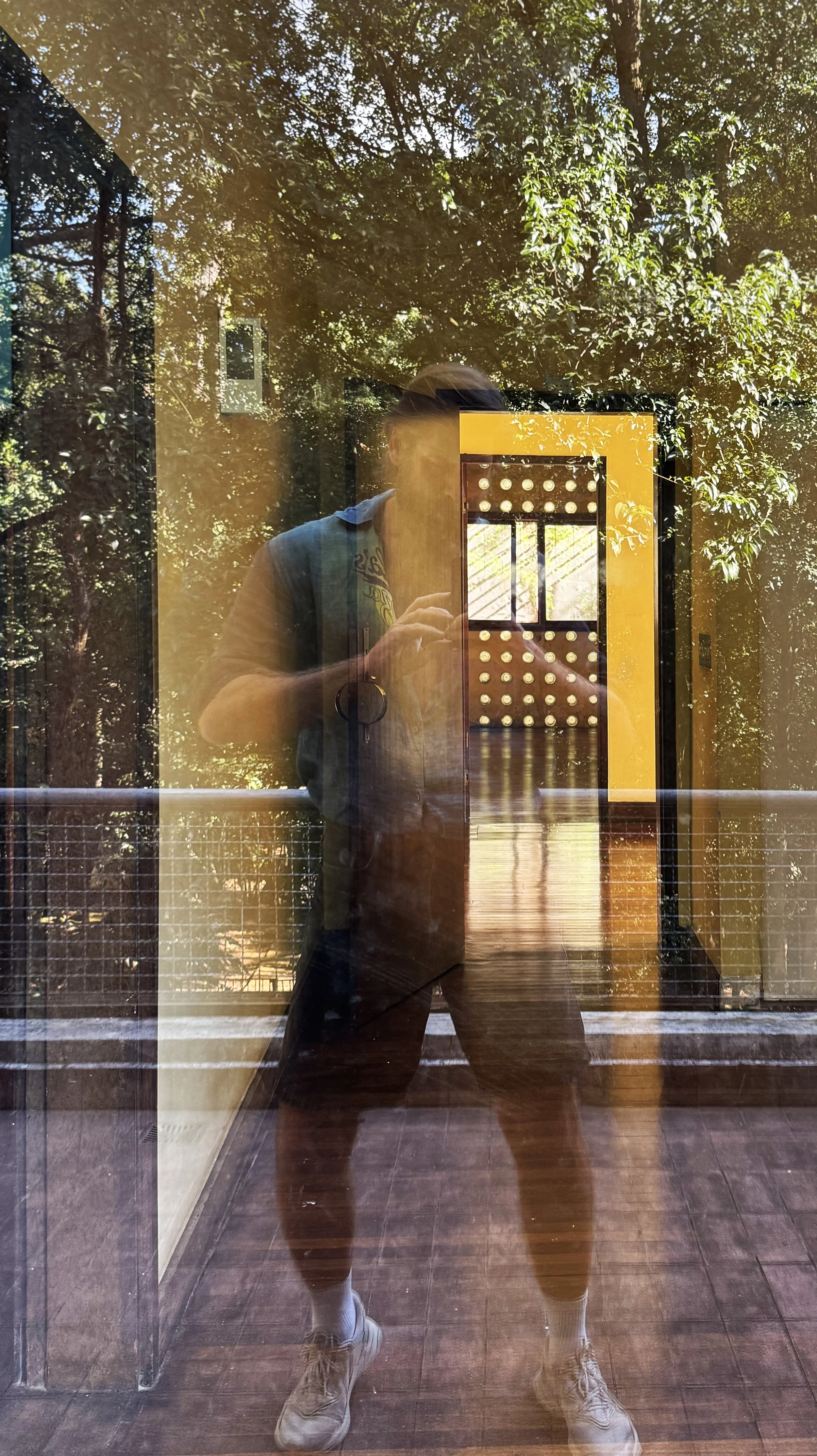 Reflection of a person taking a photo in a glass door, with trees and yellow structure in the background.