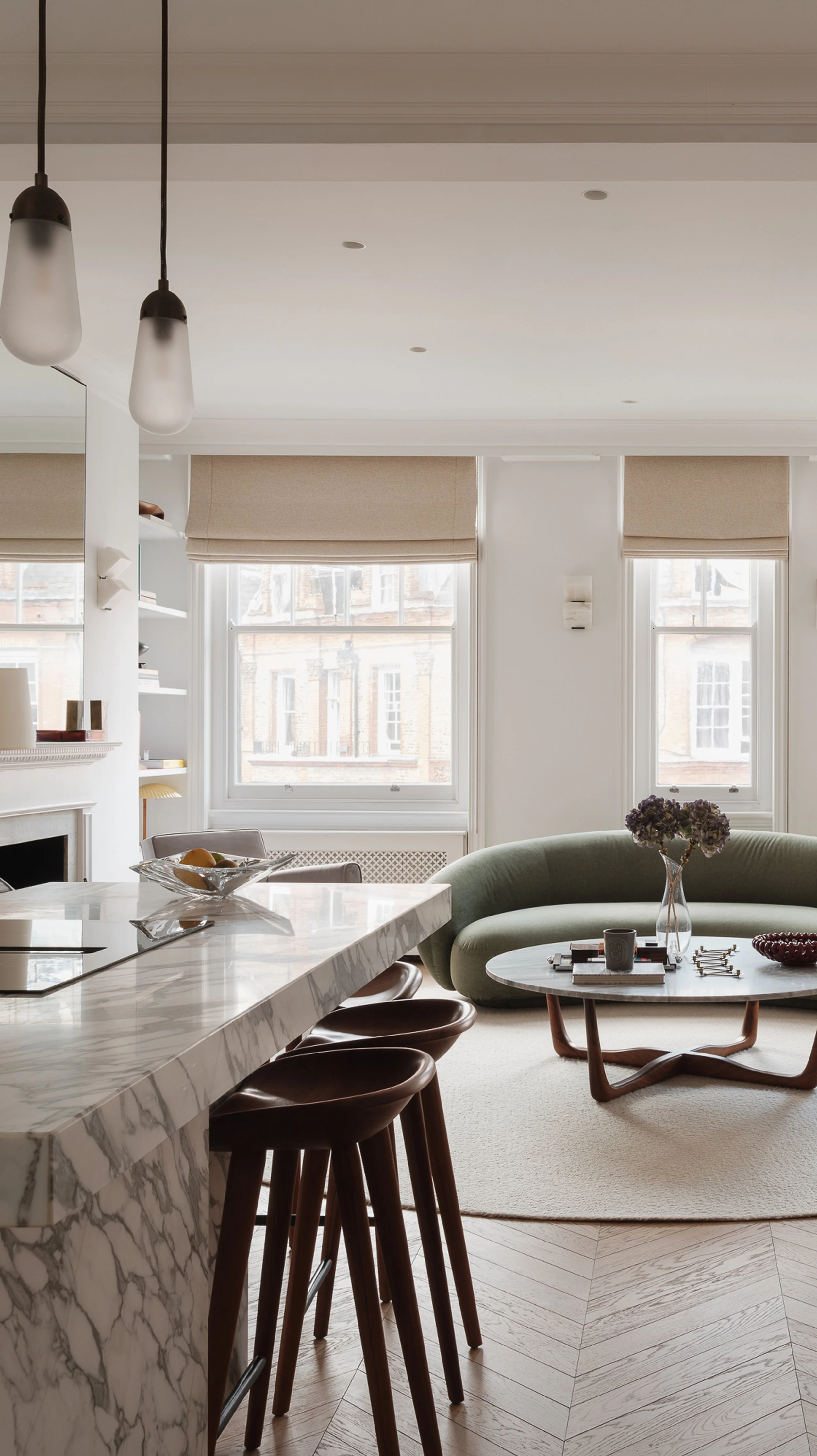 A modern living room with large windows, a marble kitchen island with three bar stools, a green curved sofa, a round coffee table with a vase of flowers, books, and decorative items.