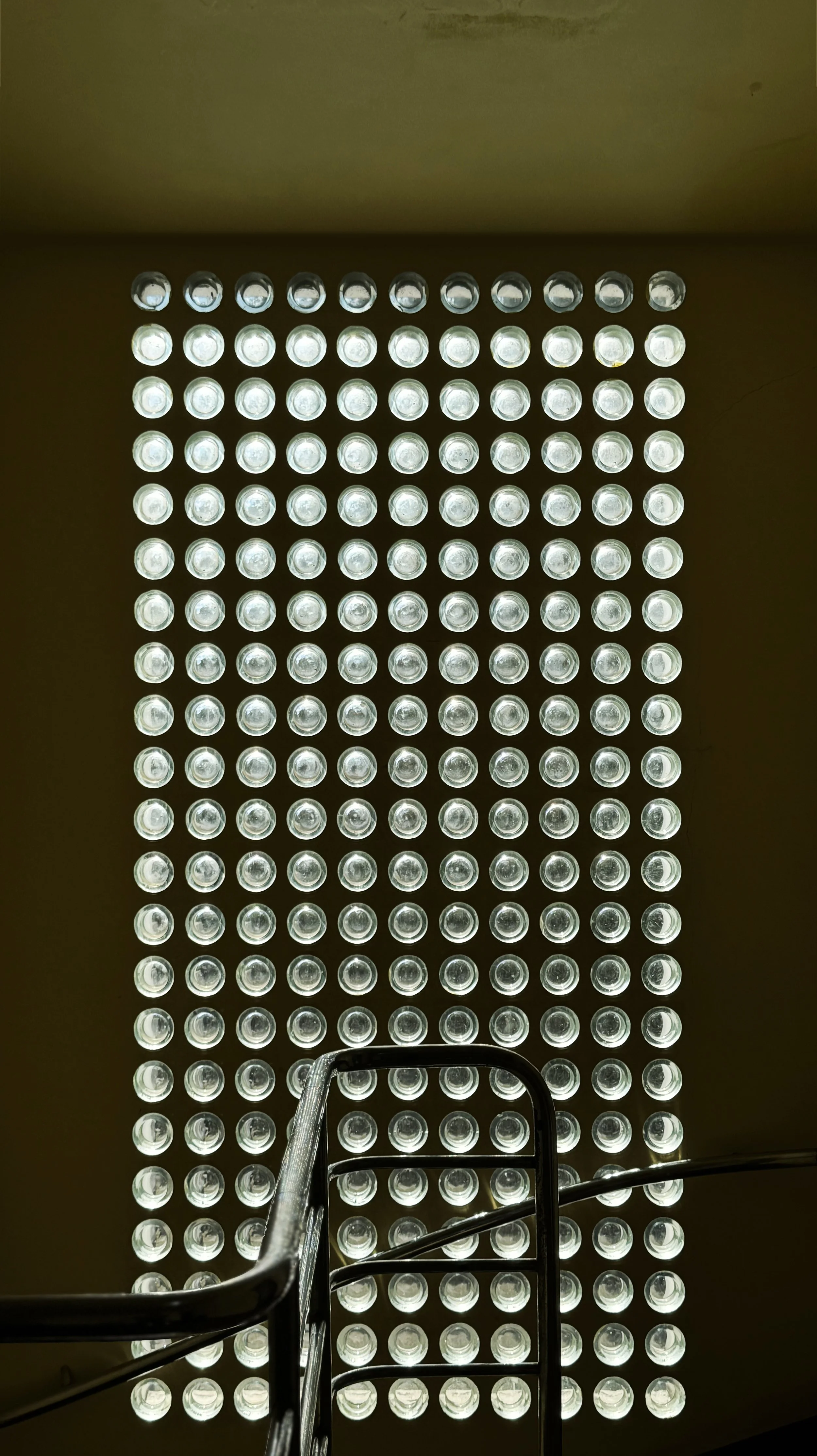 Close-up view of a wall with numerous illuminated round lights arranged in a grid pattern, with metal handrails in the foreground.