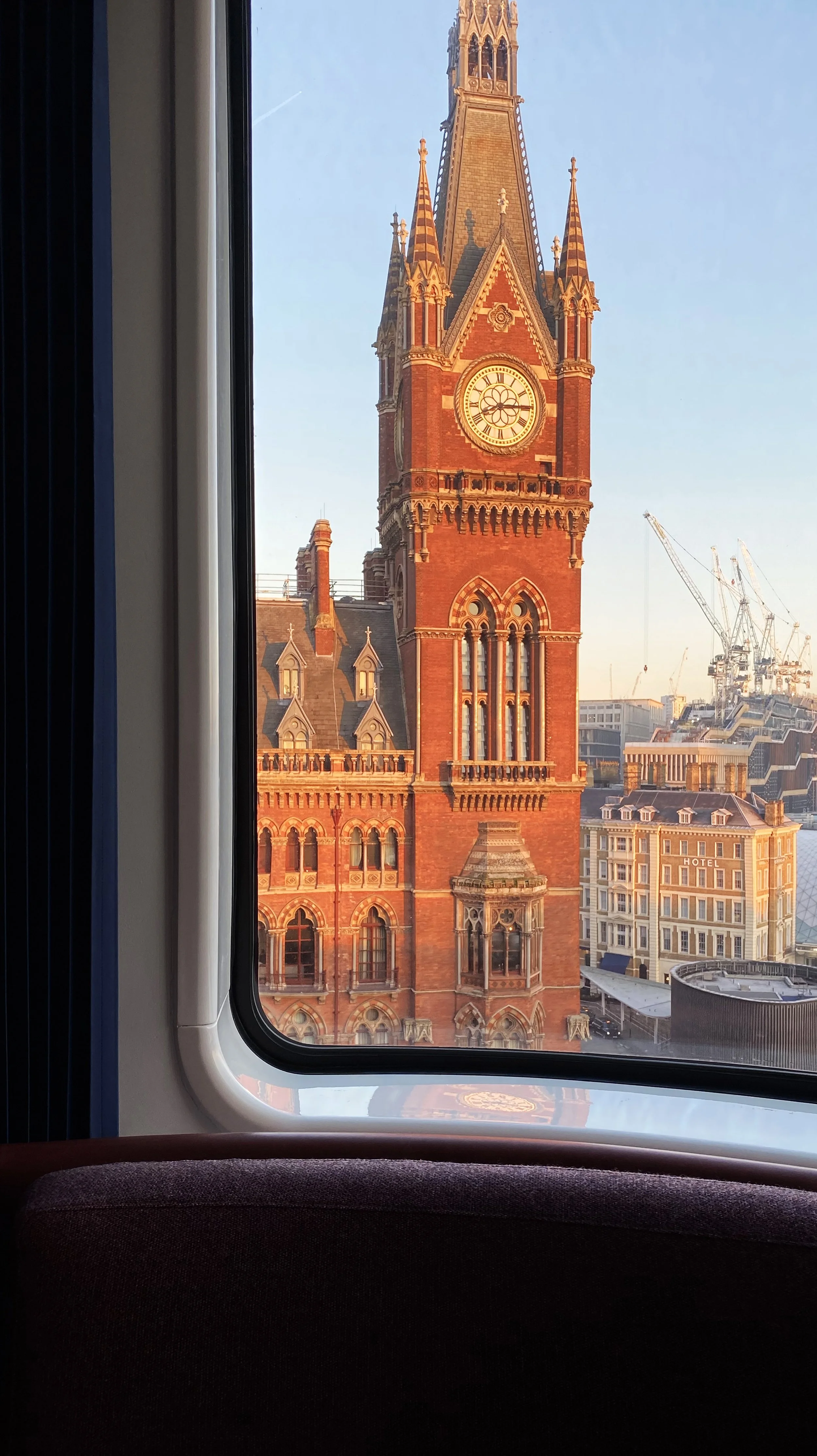 View of a red brick clock tower seen through a window from inside a building or train, with some buildings and construction cranes in the background.