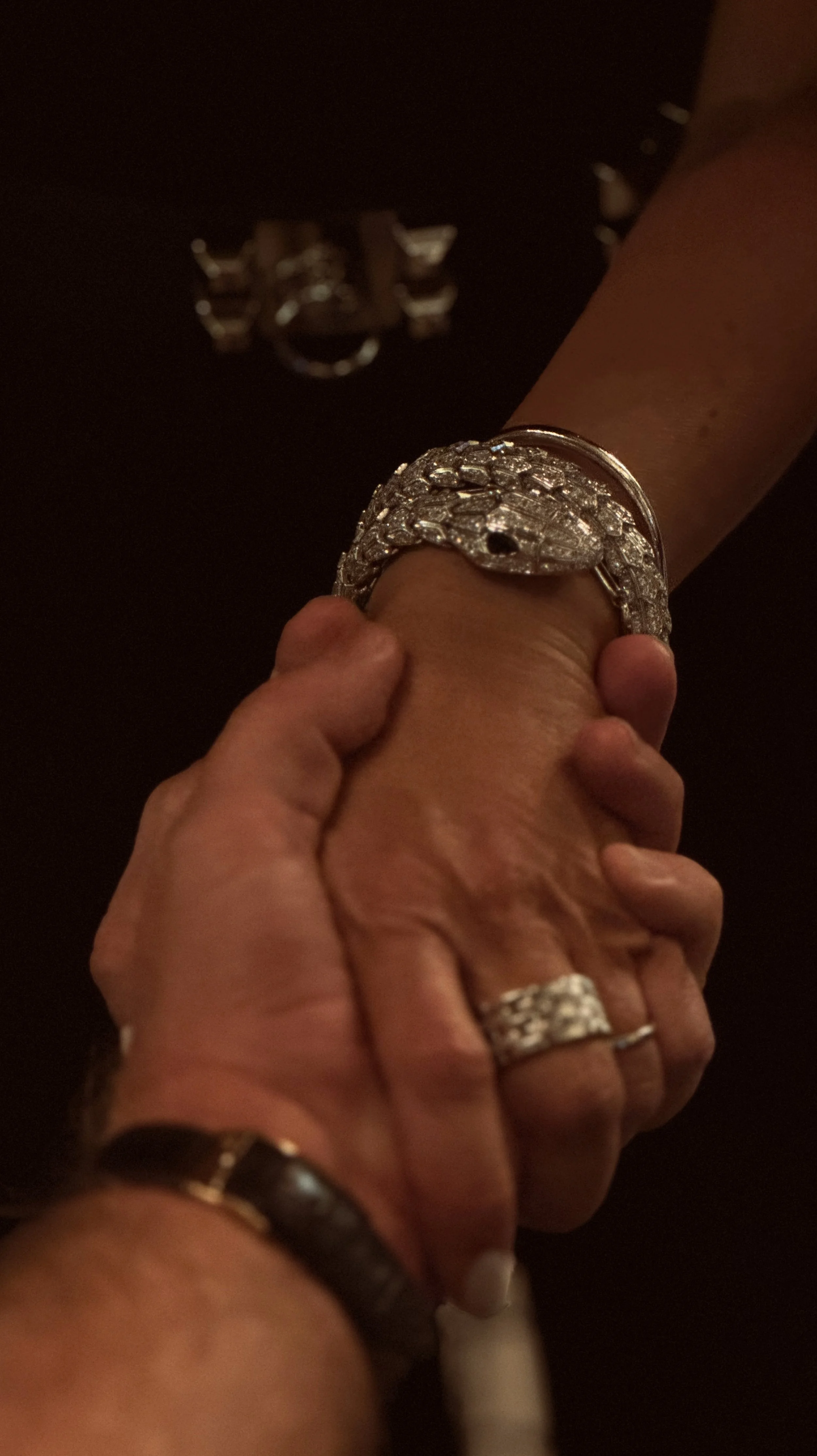 A close-up of two hands, one with a large diamond ring and palm resting on the other person's wrist, which is wearing a silver bracelet. The background is dark with a partially visible silver ring in the background.