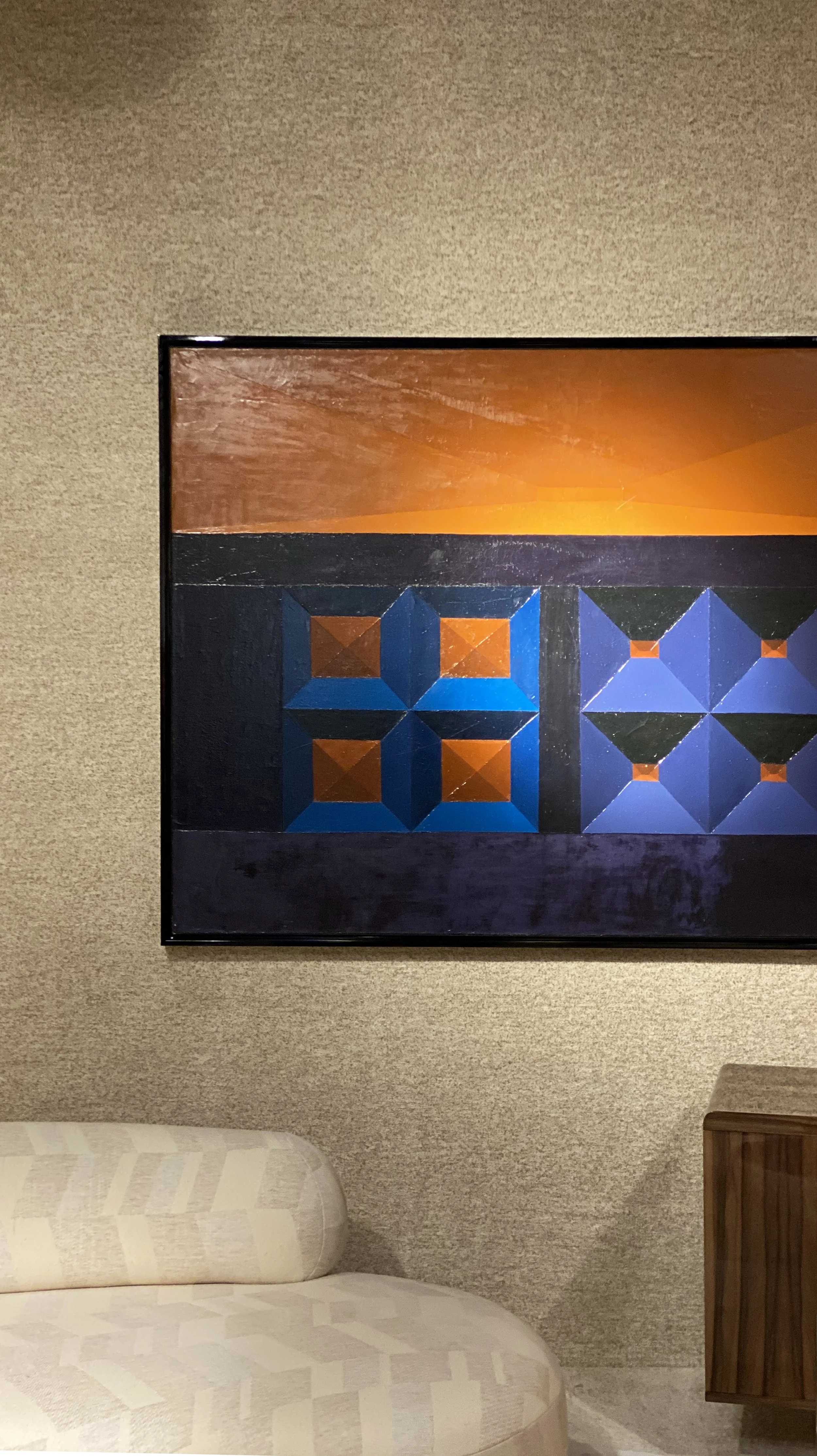 Benjamin Liatoud develops premium architecture and interior design for private clients and investors. A section of a living room wall with a large framed geometric abstract painting, a beige upholstered sofa, and a wooden side table.