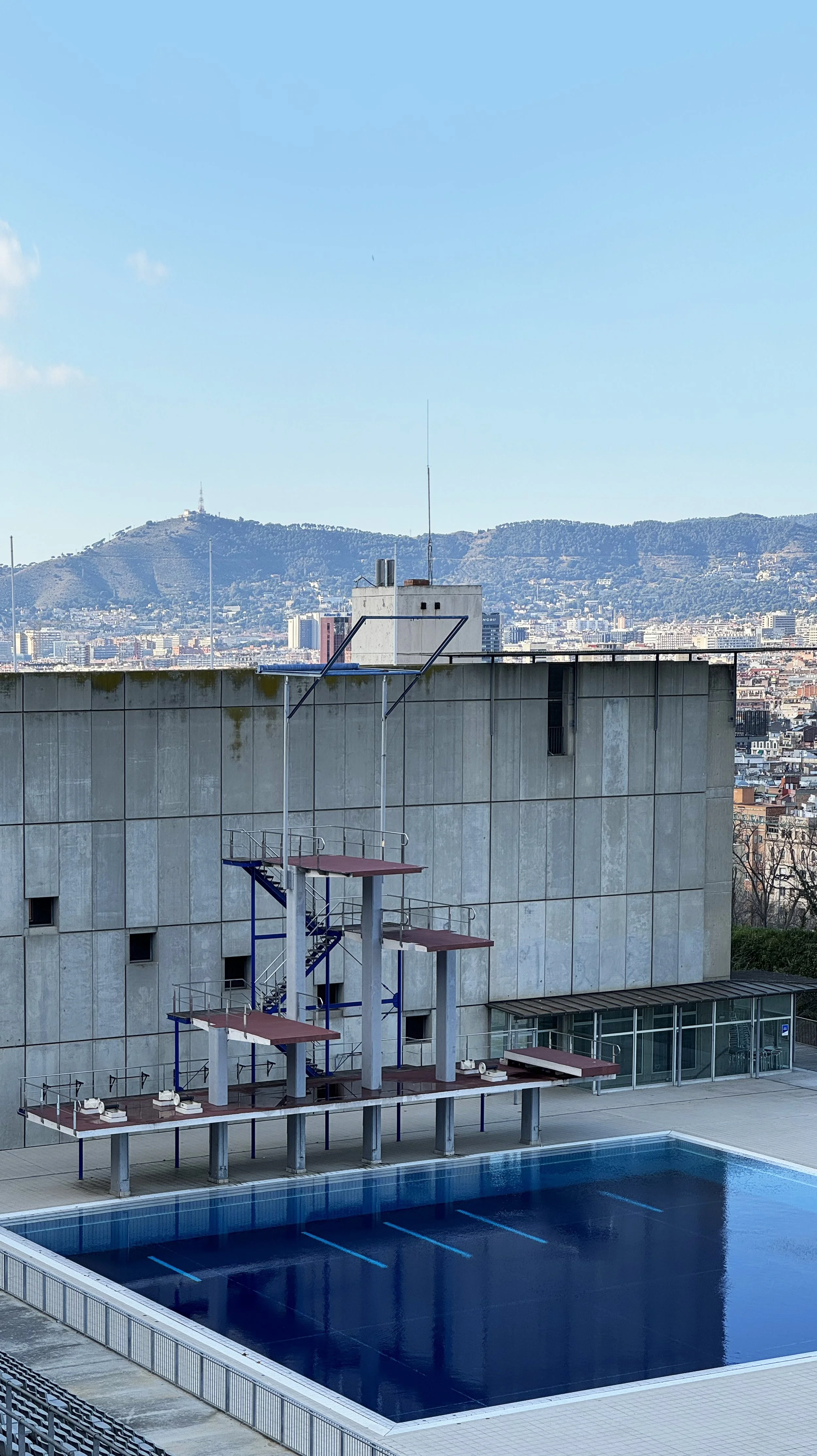 A swimming pool with diving platforms in an urban setting with mountainous hills in the background.