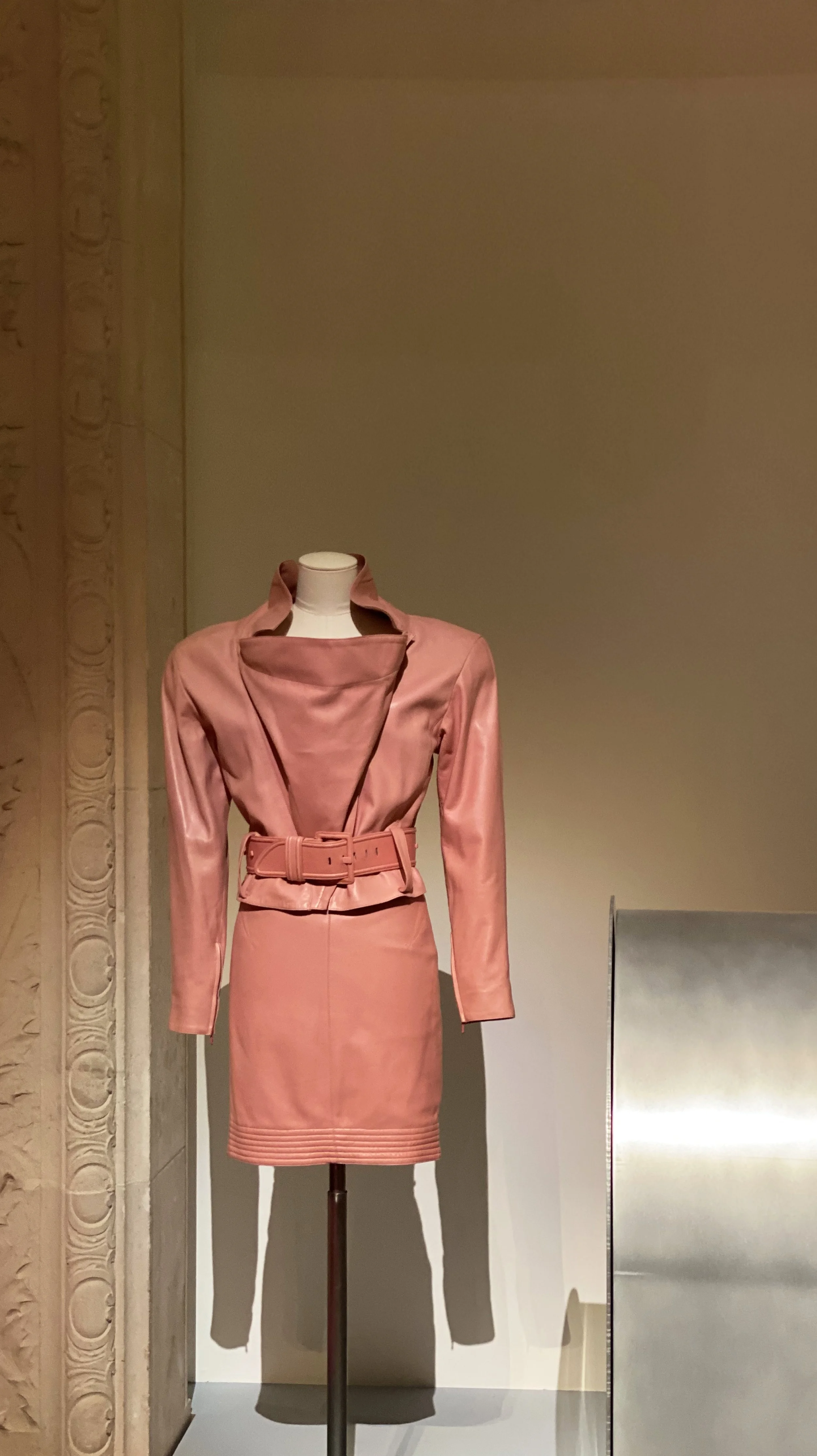 Pink satin dress with a matching belt and wide collar displayed on a mannequin in a museum.