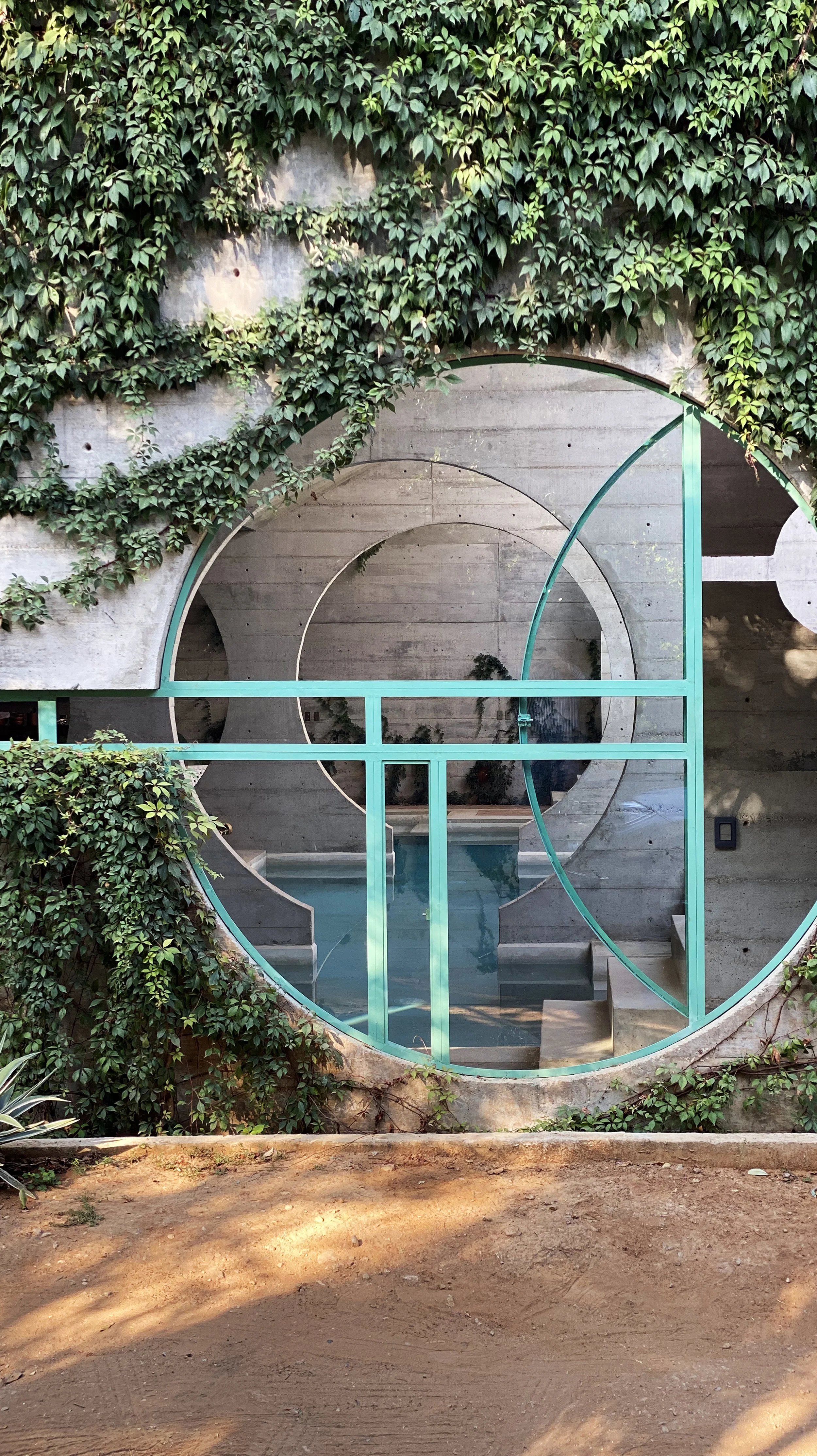 Modern concrete structure with large circular windows framed in aqua, greenery covering parts of the structure, a small pool or water feature inside, and a dirt foreground.