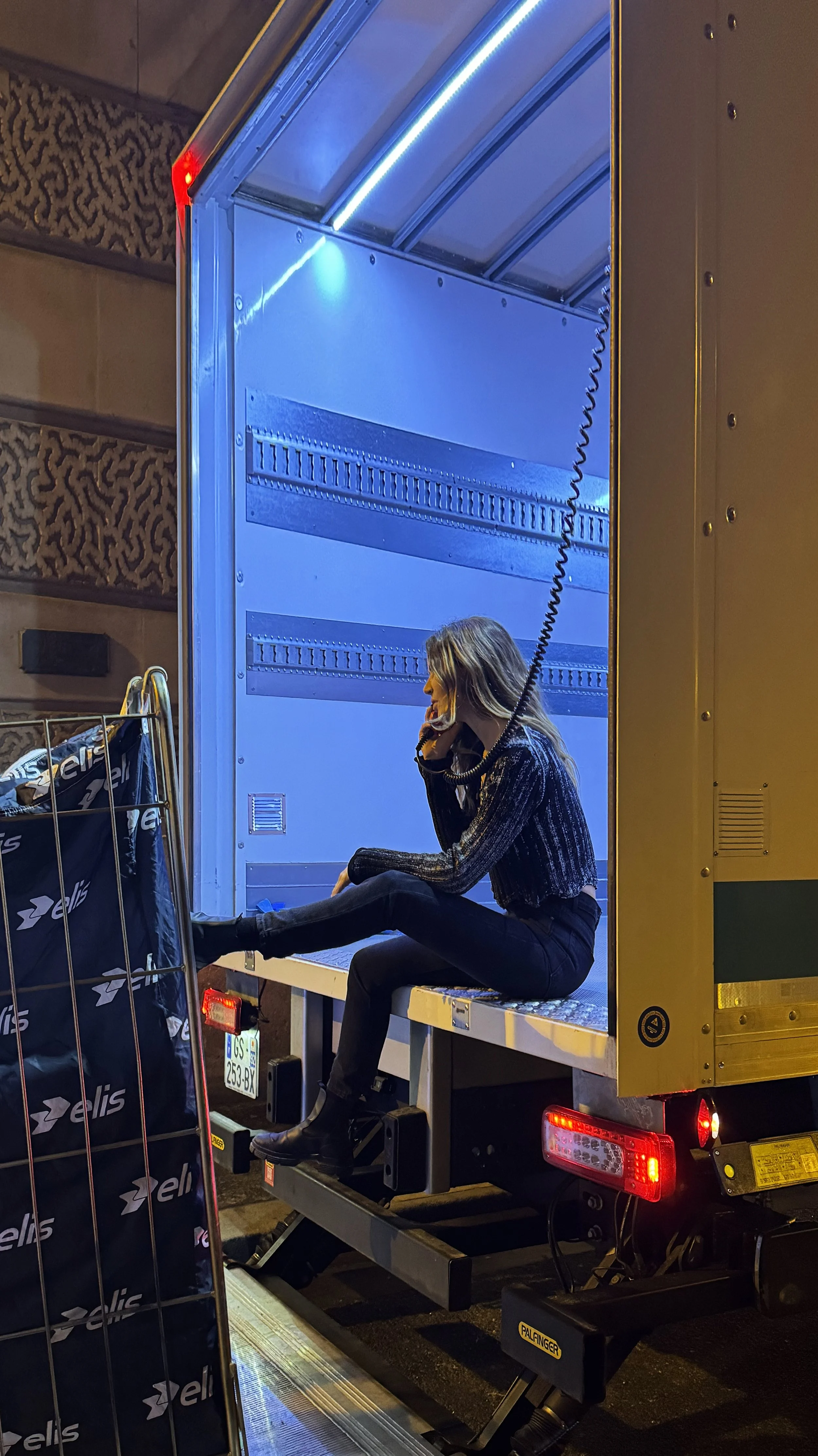 A woman sitting on the edge of a truck bed, talking on a landline phone, with her legs crossed, illuminated by blue lighting, and surrounded by shopping bags.