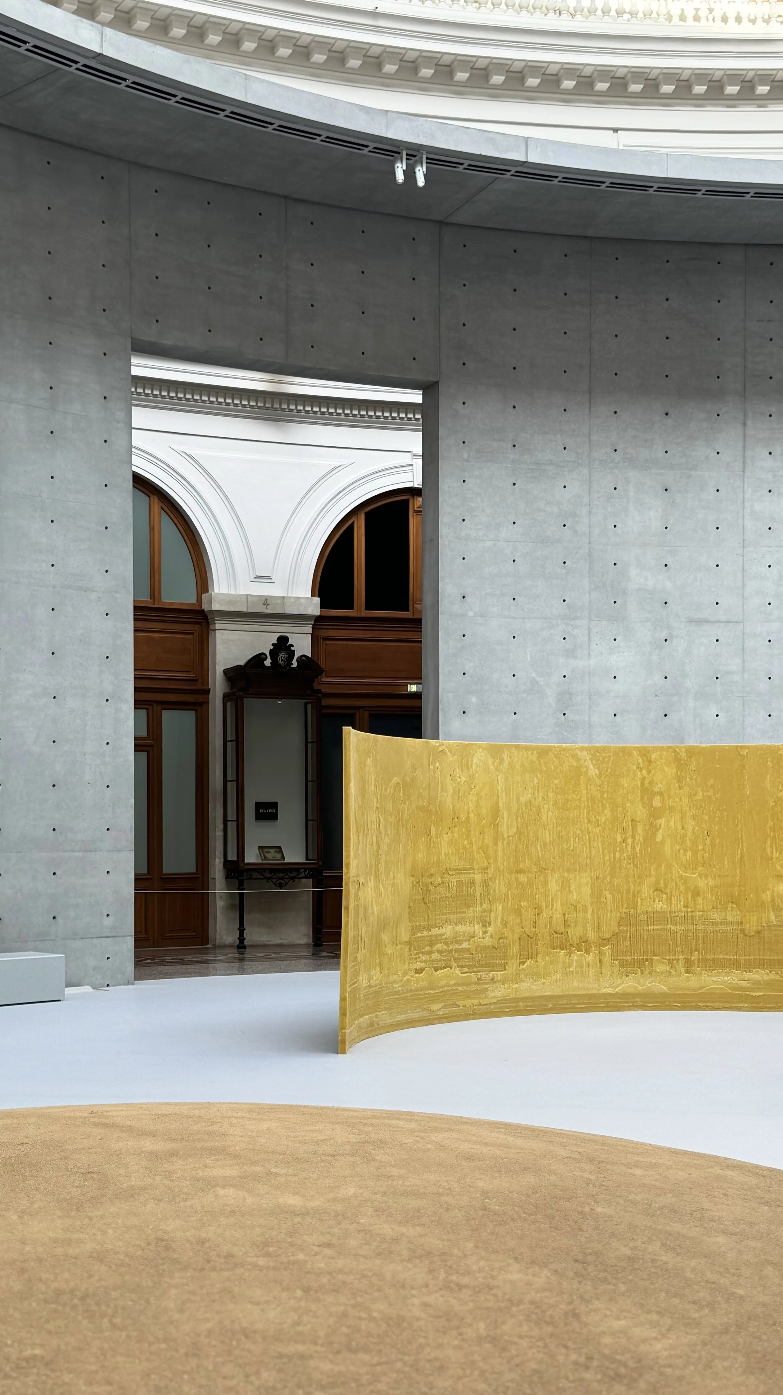 Interior of a modern museum with concrete walls, a curved yellow divider, and classical architectural details visible through an opening in the wall, including arched windows with wooden frames and ornate white molding.