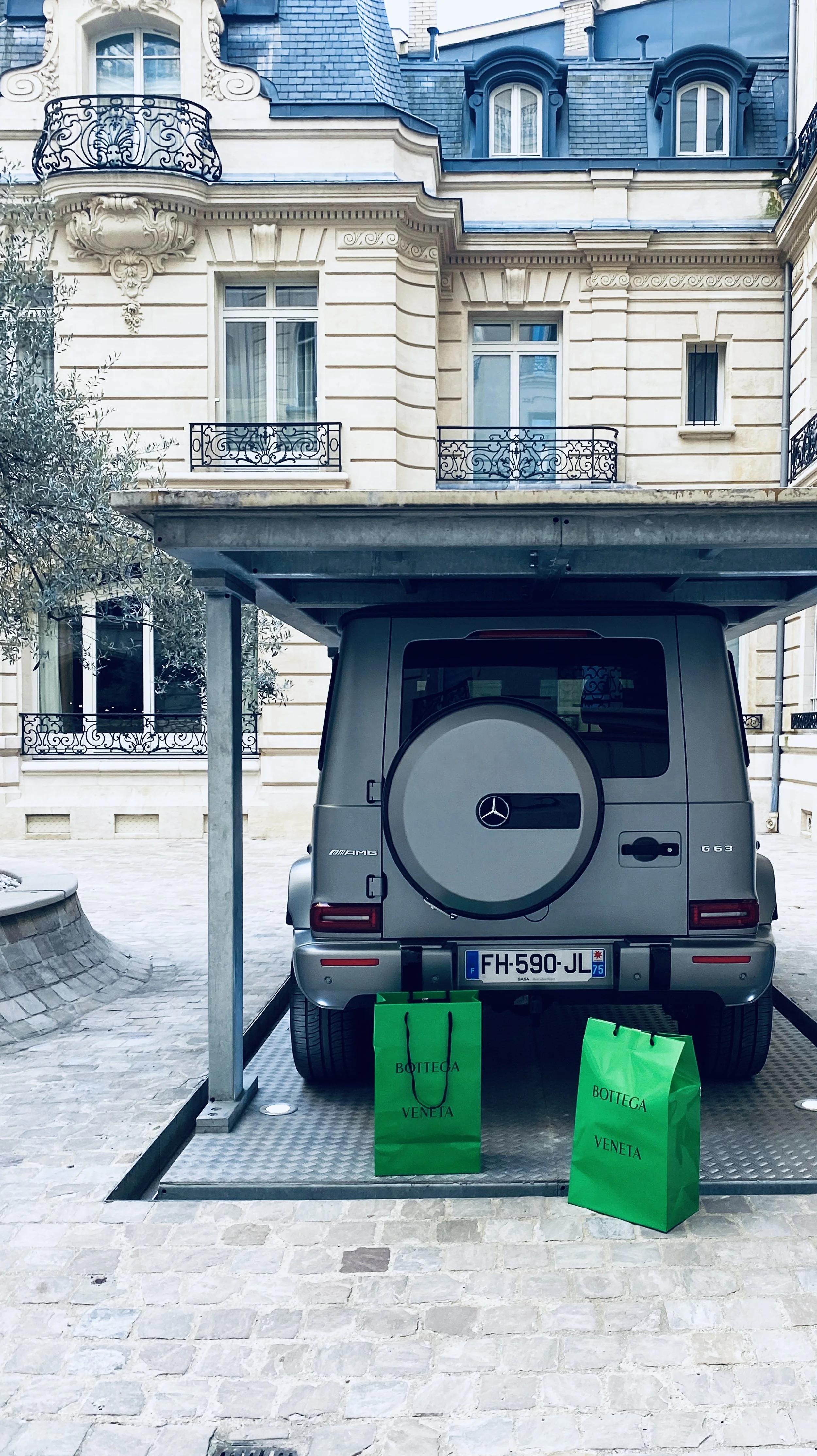 A gray Mercedes-Benz G-Class SUV parked under a carport in front of a beige European-style building with ornate architectural details and black wrought iron balconies. Two bright green shopping bags labeled "Bottega Veneta" sit on the ground behind t