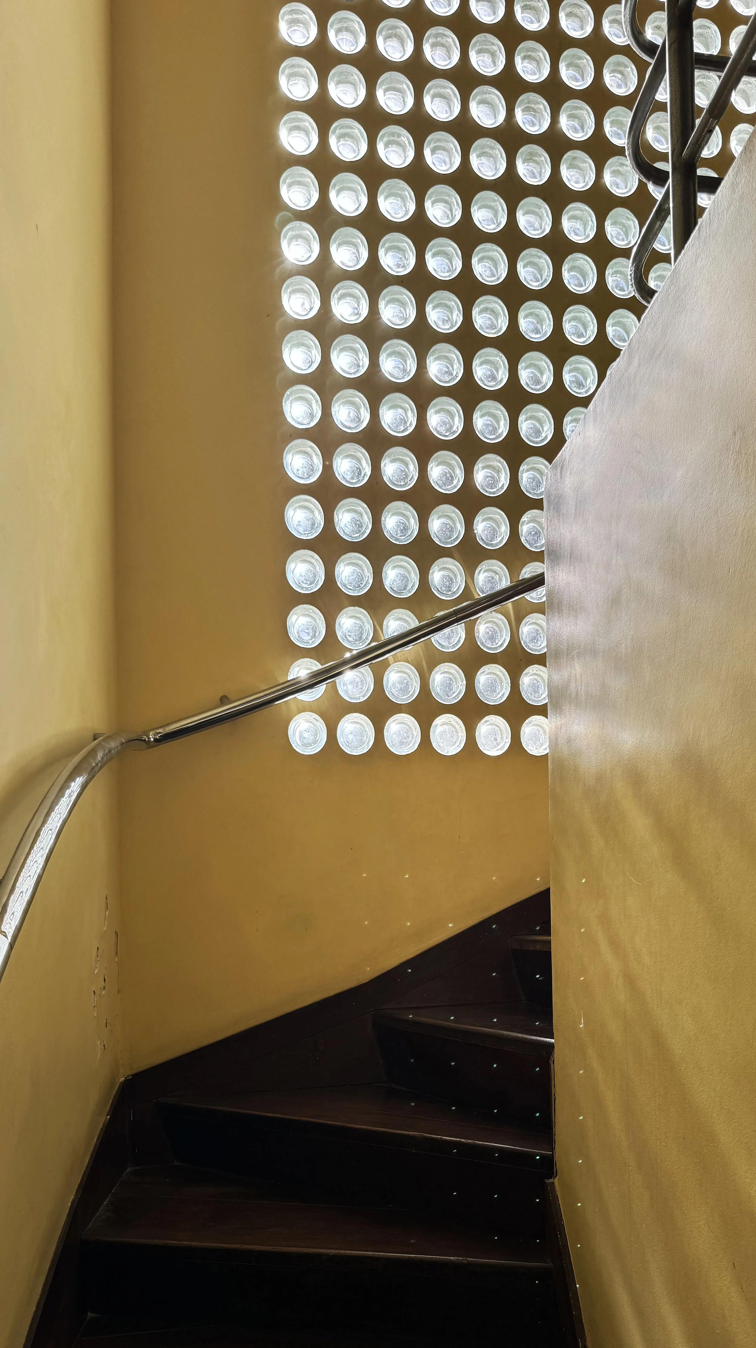 Indoor staircase with dark wooden steps, a metal handrail, yellow walls, and a decorative wall with circular glass blocks allowing light through.