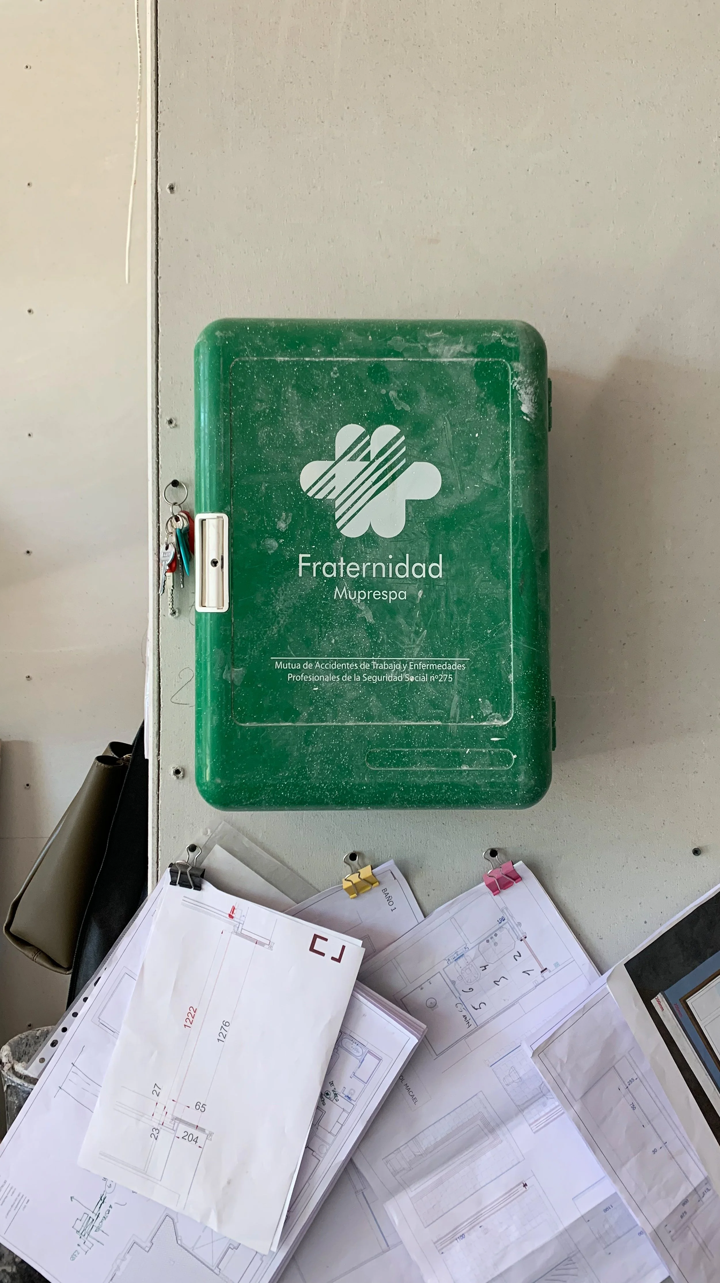 Green first aid kit labeled "Fraternidad Muprespa" attached to a wall with some documents and keys hanging nearby.