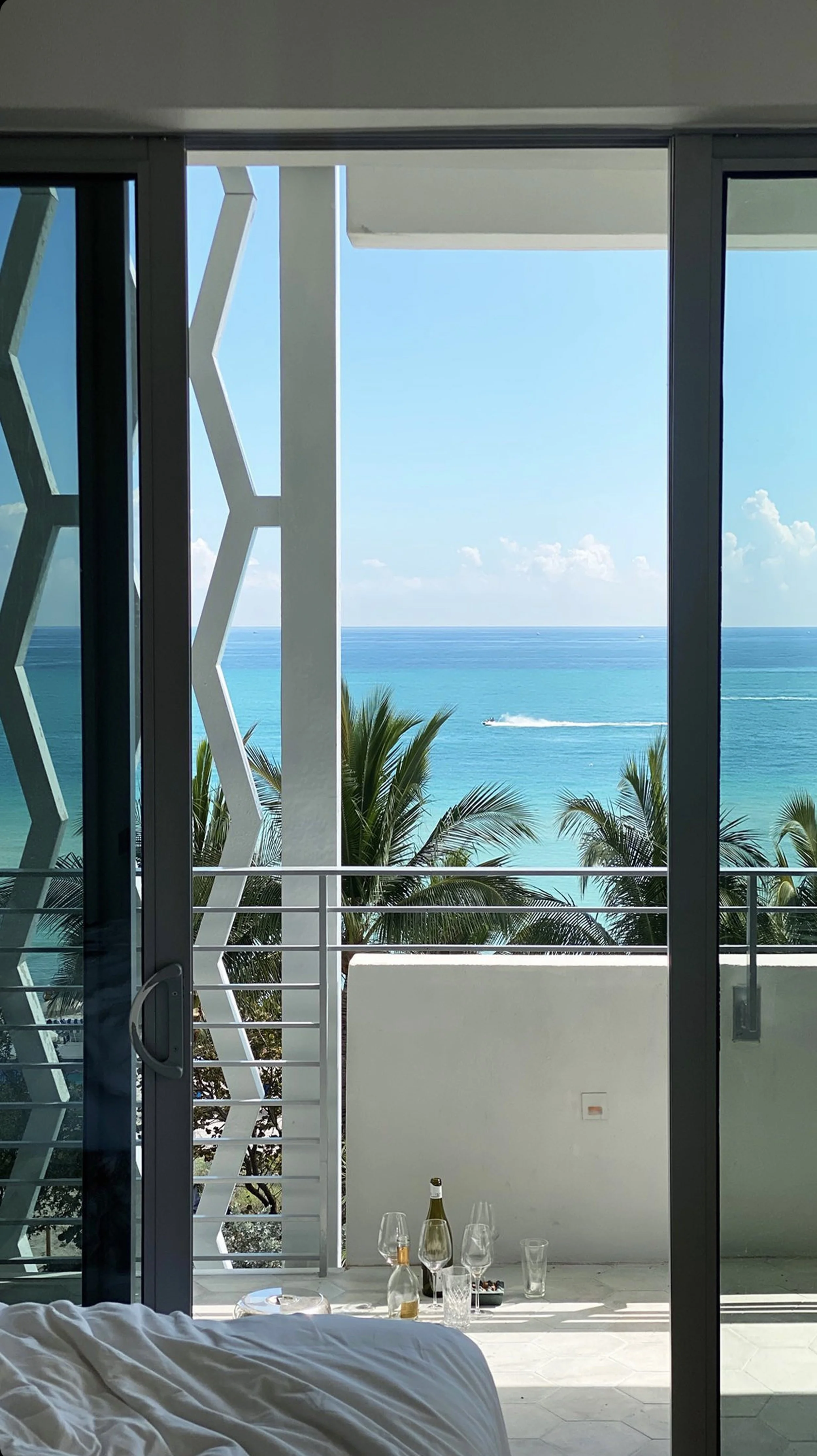 Hotel room balcony with glass sliding door, overlooking palm trees and the ocean with a boat in the distance, and a small table with wine bottles, glasses, and snacks.