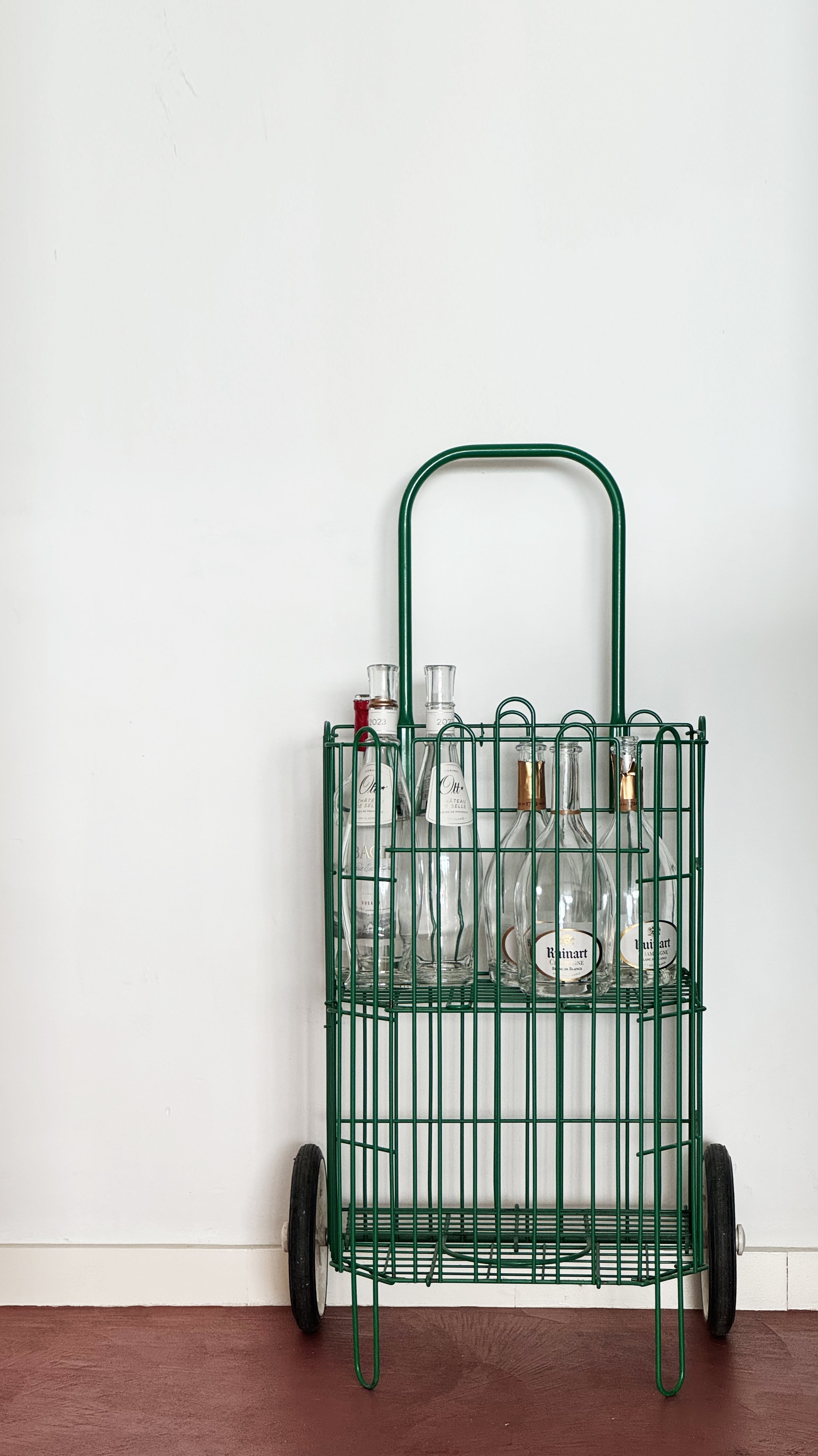 Green metal cart with wheels holding empty glass bottles, set against a white wall and red wooden floor.
