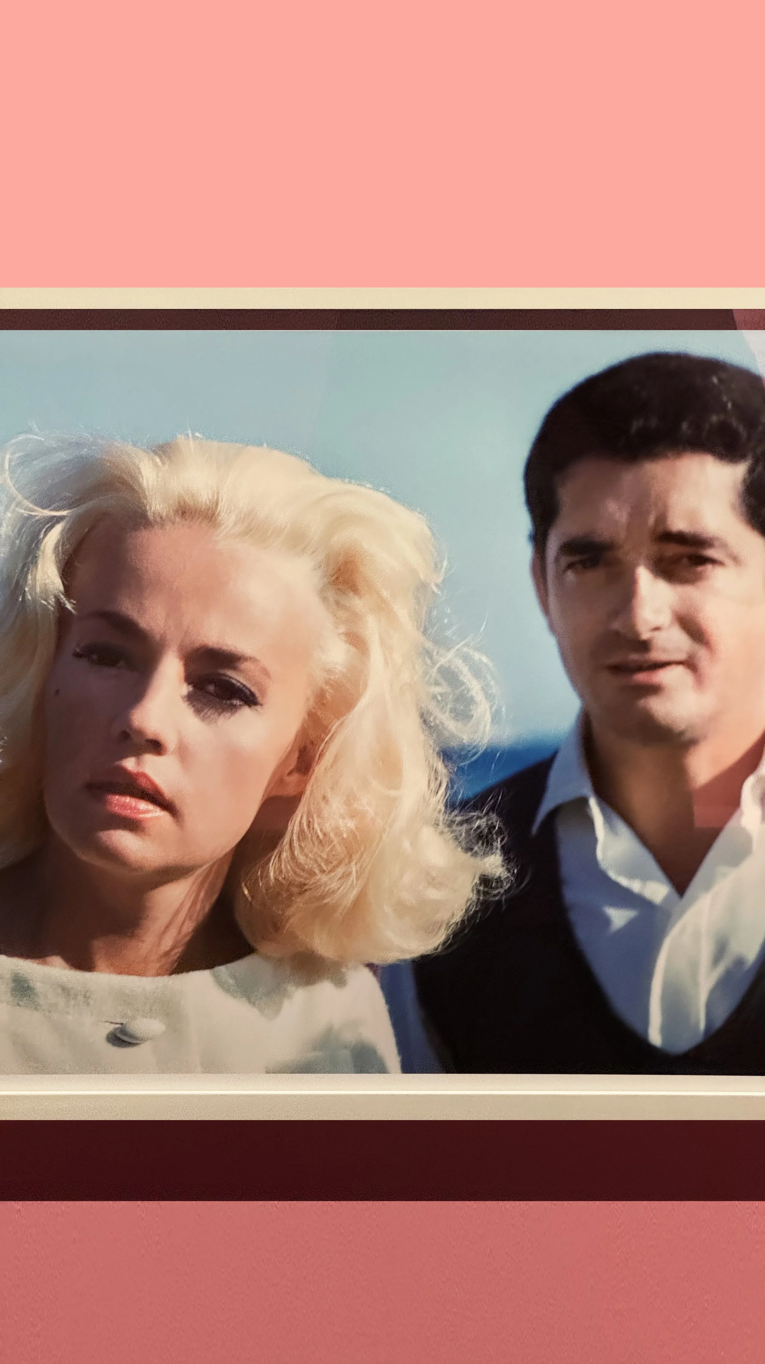 A photograph of a woman with platinum blonde hair and a man with dark hair, both dressed in semi-formal attire, outdoors with a clear blue sky in the background.