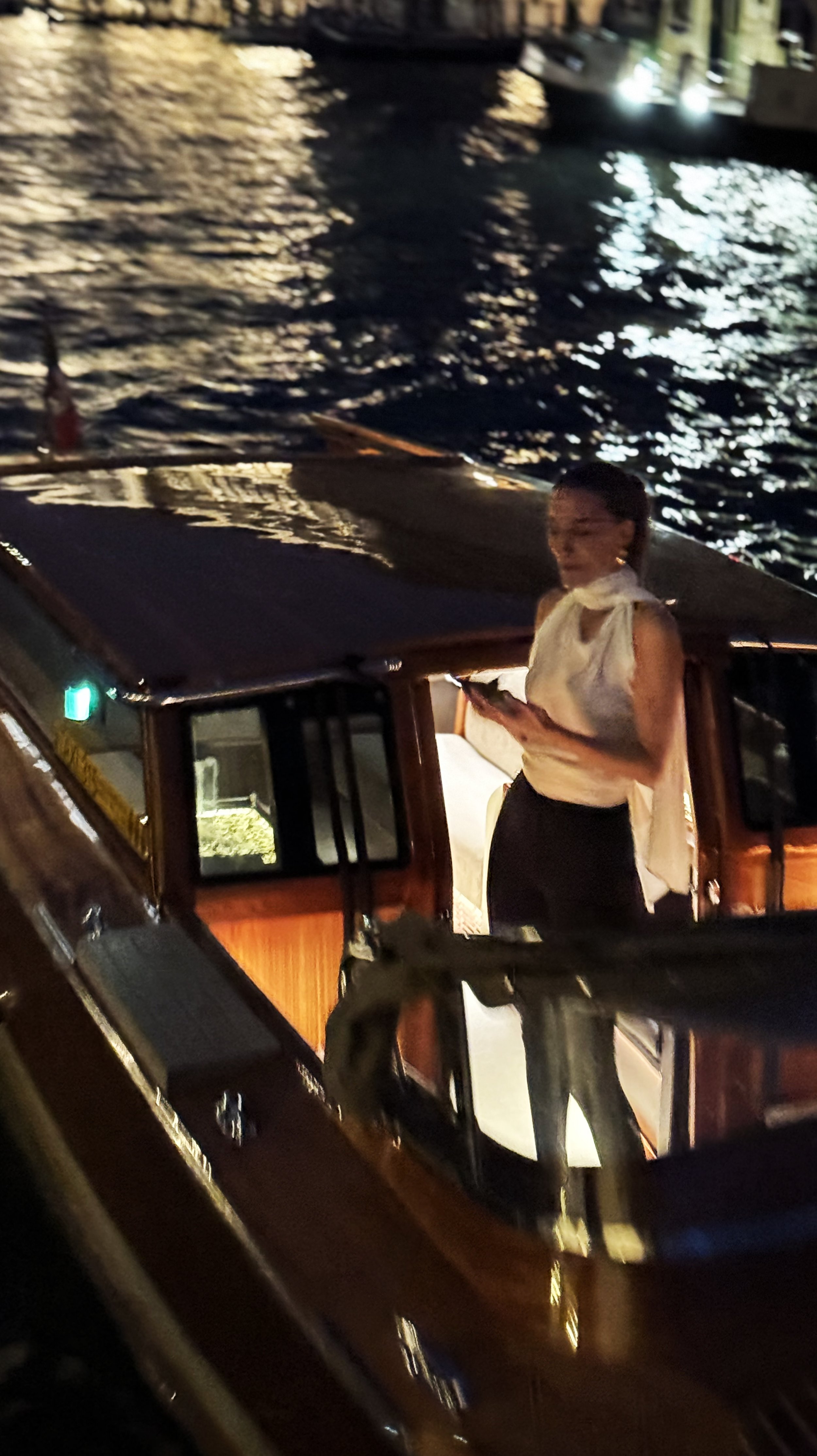 A woman standing on a boat at night, near a body of water with reflections of lights.