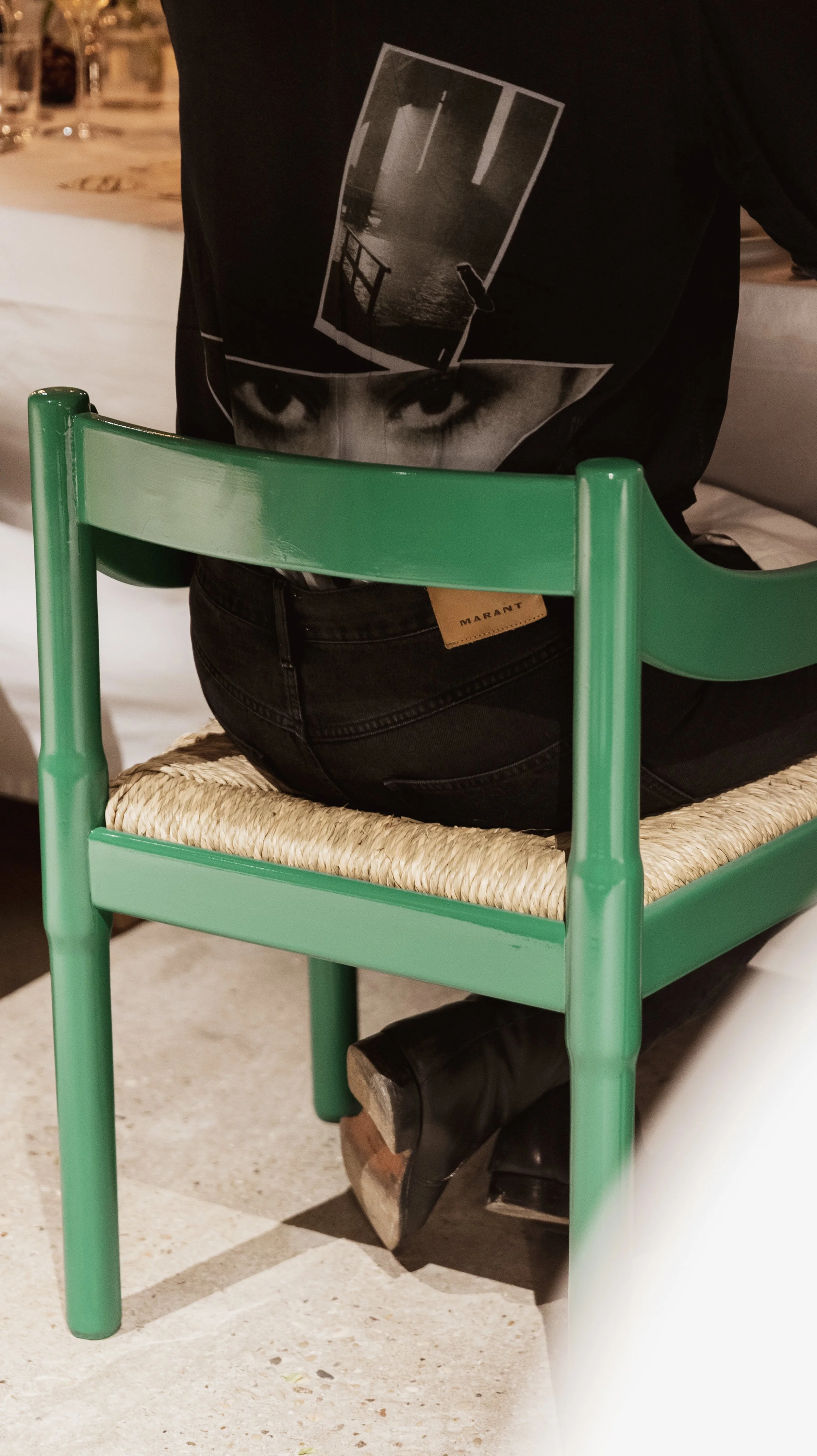 Person sitting on a green chair, wearing black jeans and black boots, with a black t-shirt that has a graphic design on the back, in a well-lit indoor setting.