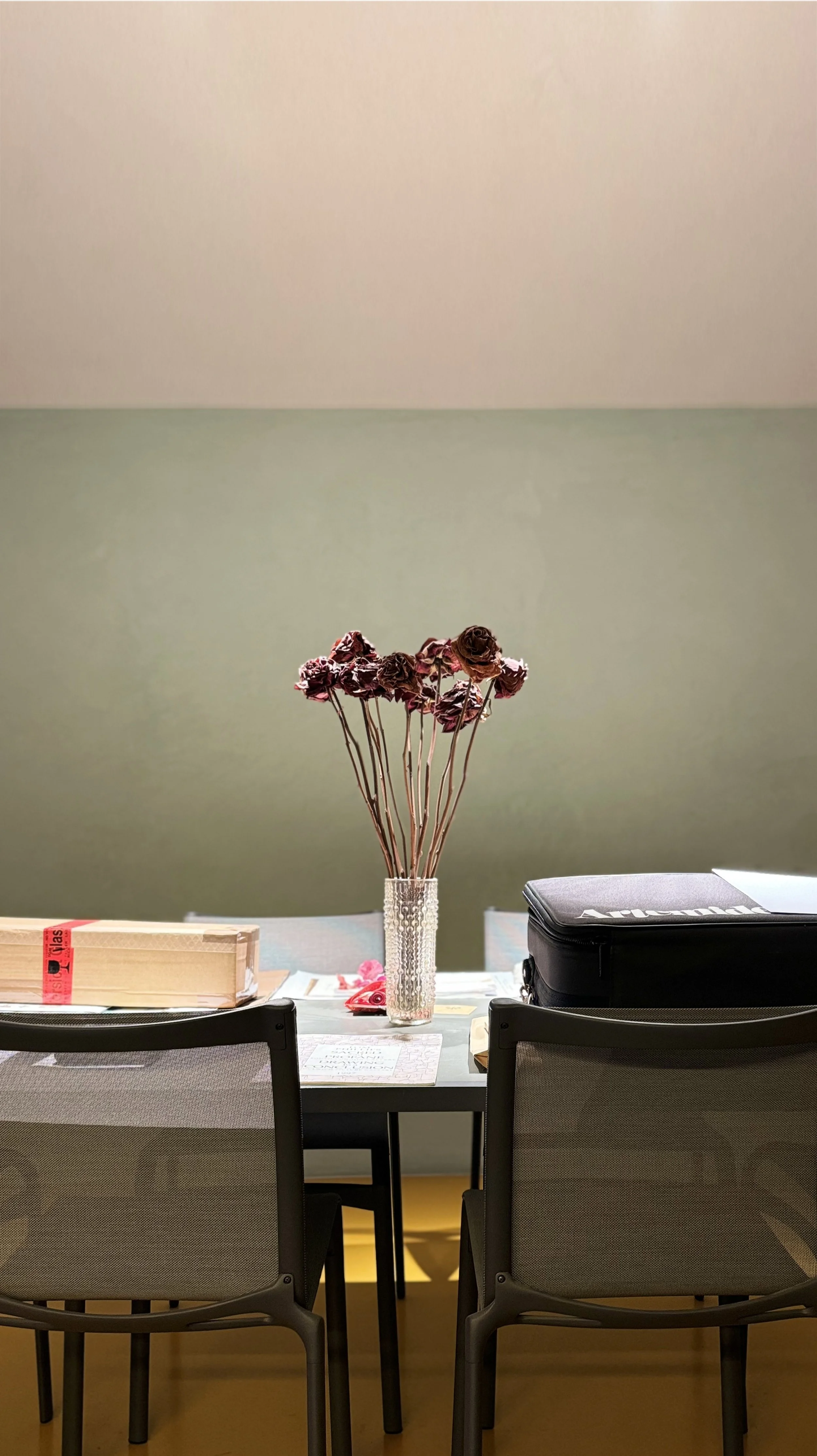 A table with two chairs, a vase with dried roses, and a closed laptop, set against a two-tone wall with light green on the bottom and beige on top.