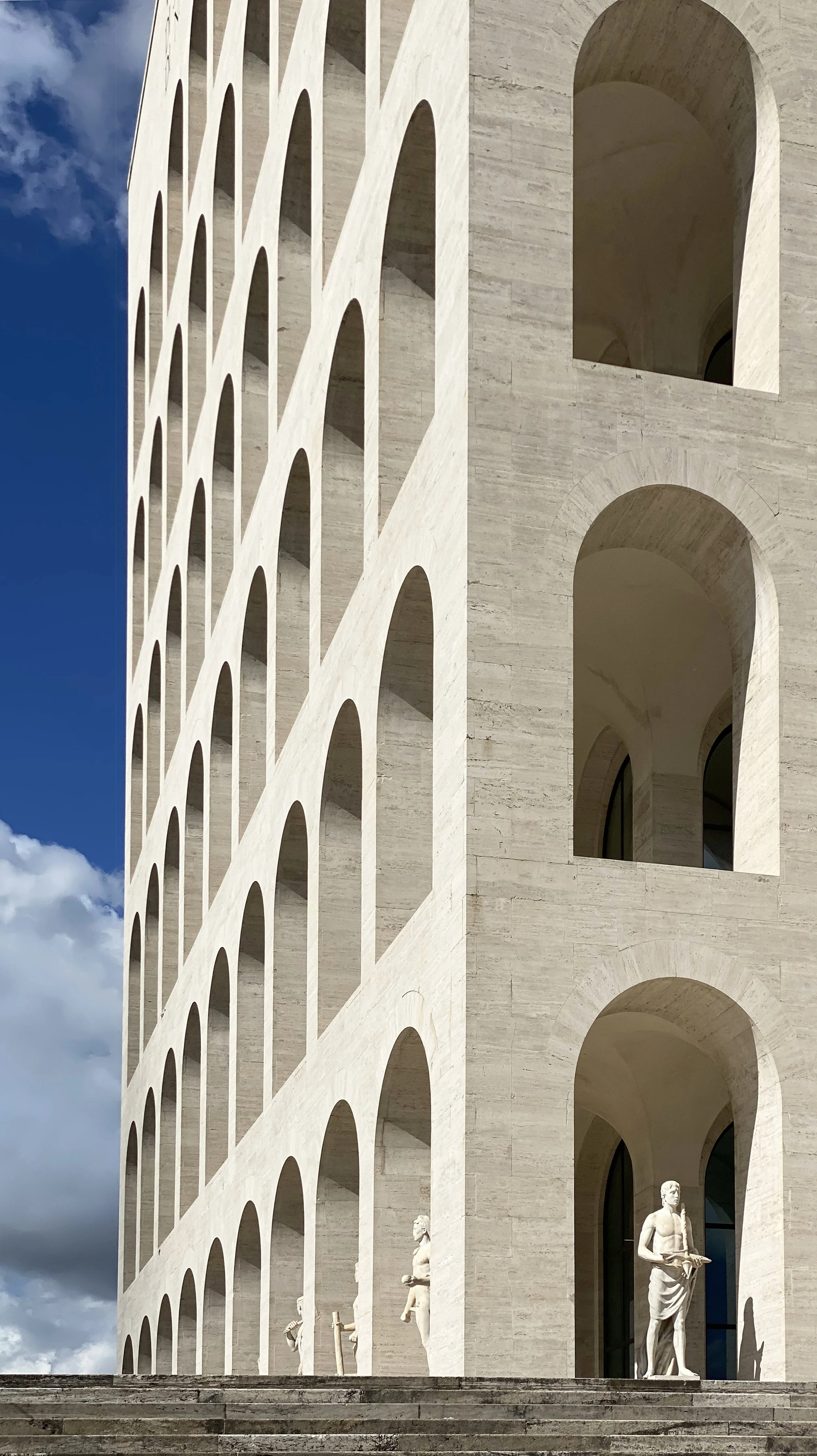 Side view of a large, modern building with multiple levels and arched openings, with white statues near the entrance and steps leading up to it, under a partly cloudy sky.