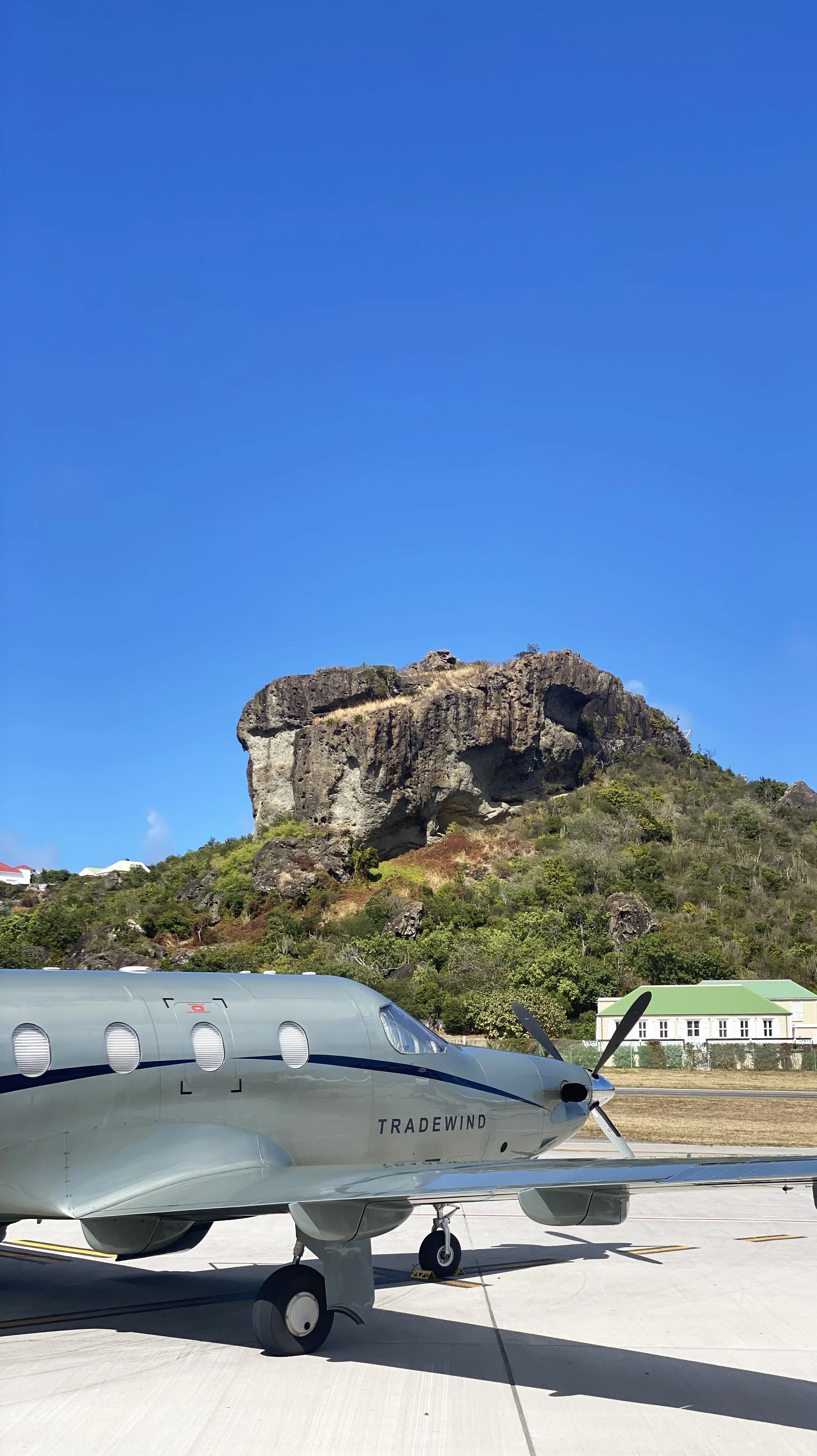 A modern airplane parked on a tarmac with a large rocky hill and green vegetation in the background under a clear blue sky.