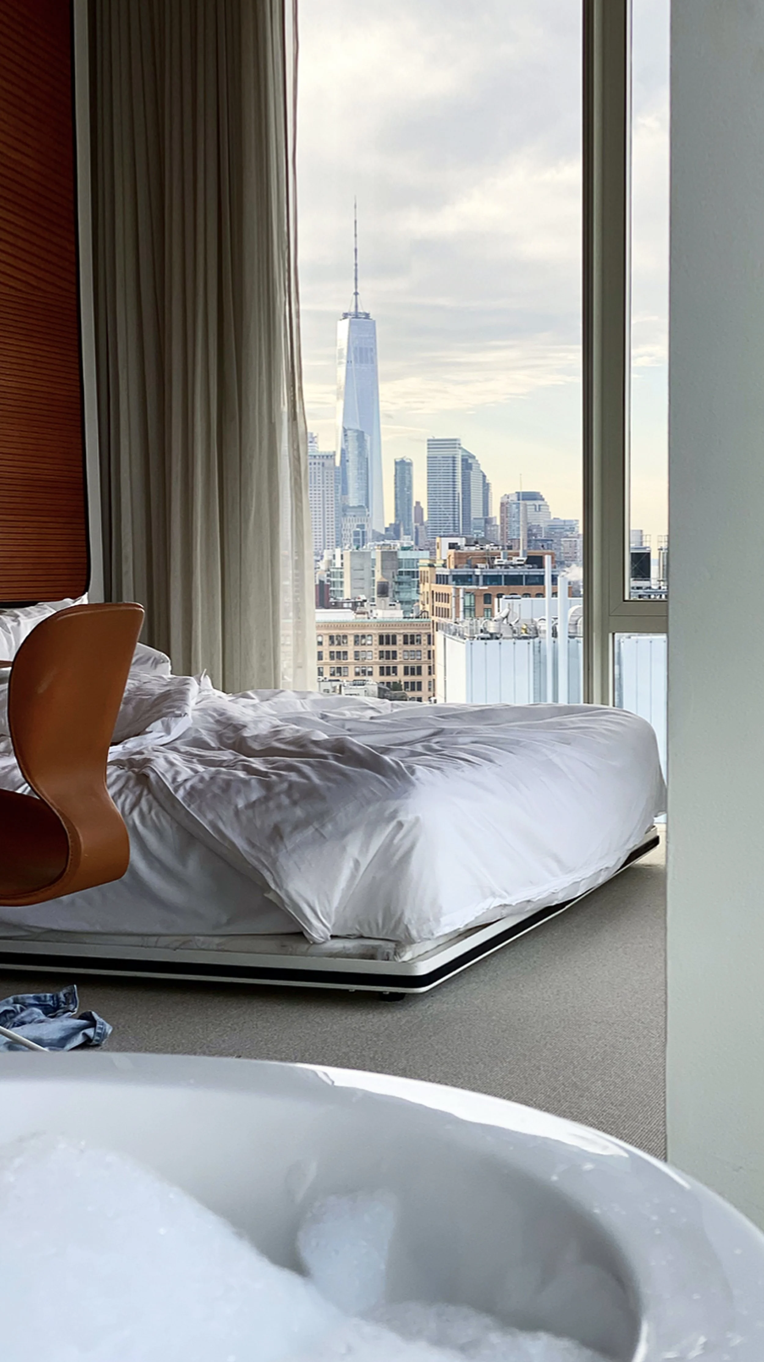 A hotel room with a bed, orange chair, and large window showing a city skyline including the One World Trade Center in New York City.