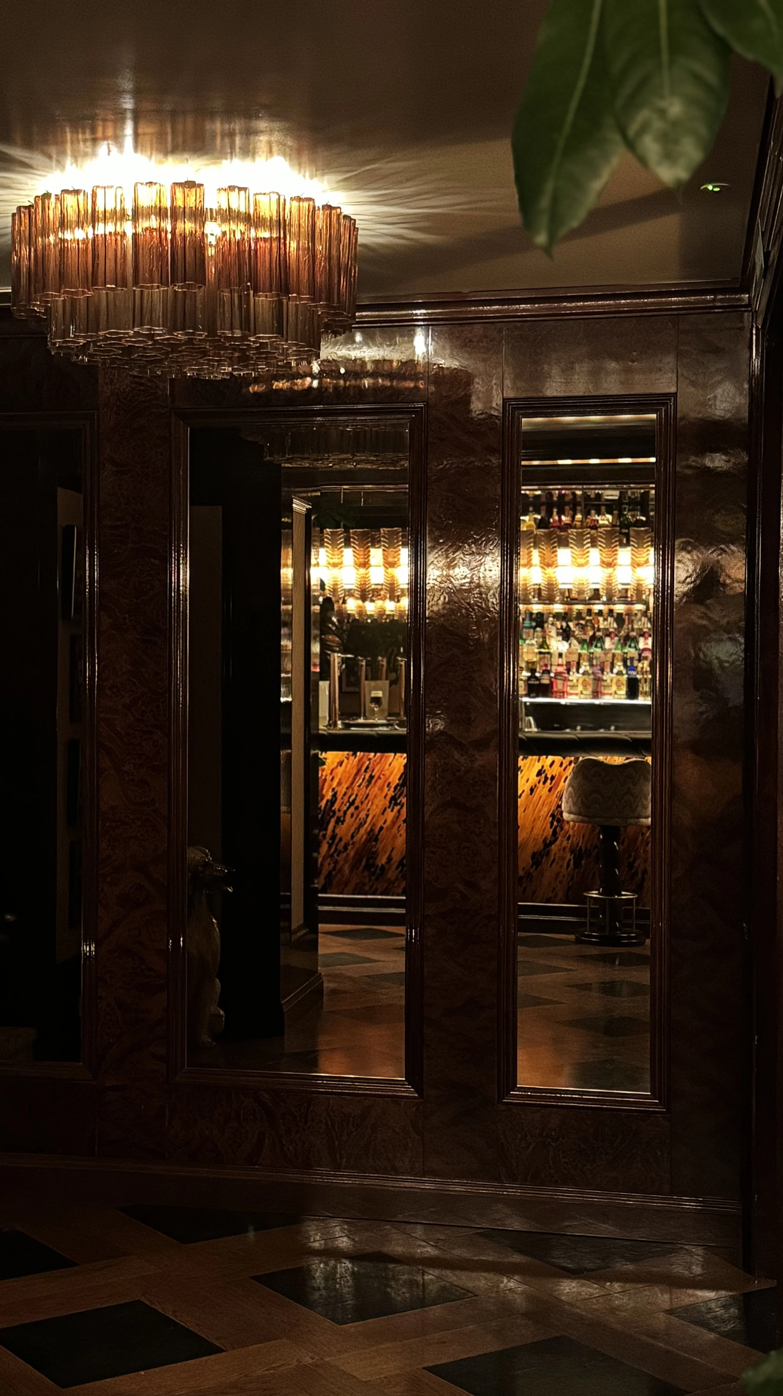 A dimly lit bar area with a backlit display of liquor bottles, a bar stool, and reflections in mirrors on the wall.